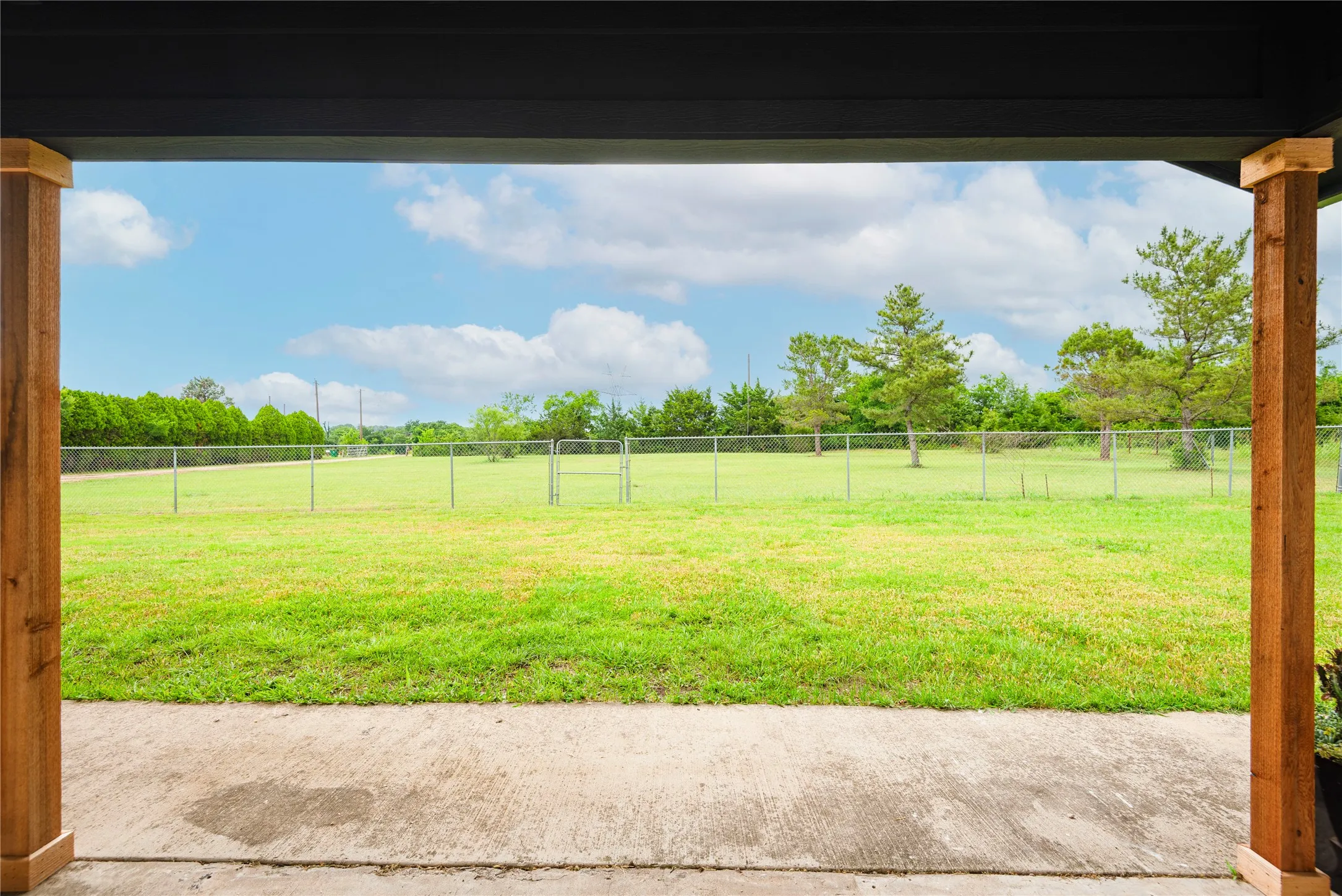 Covered concrete patio overlooking a vast grassy area with chain-link fencing, bordered by mature trees