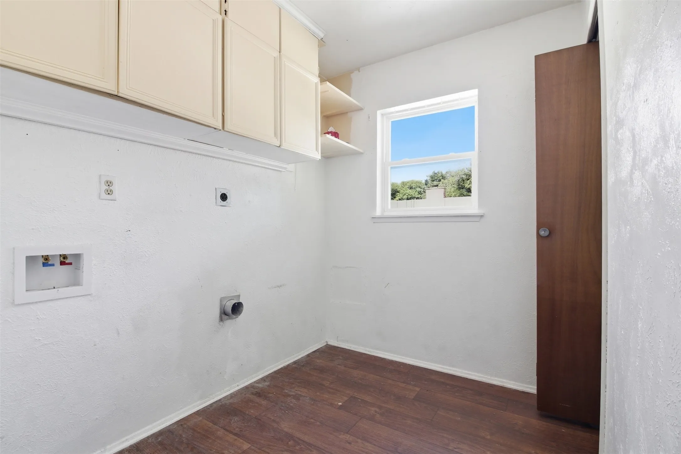 Dedicated laundry space featuring wood-finish flooring, upper cabinetry, an integrated window, and a built-in shelf