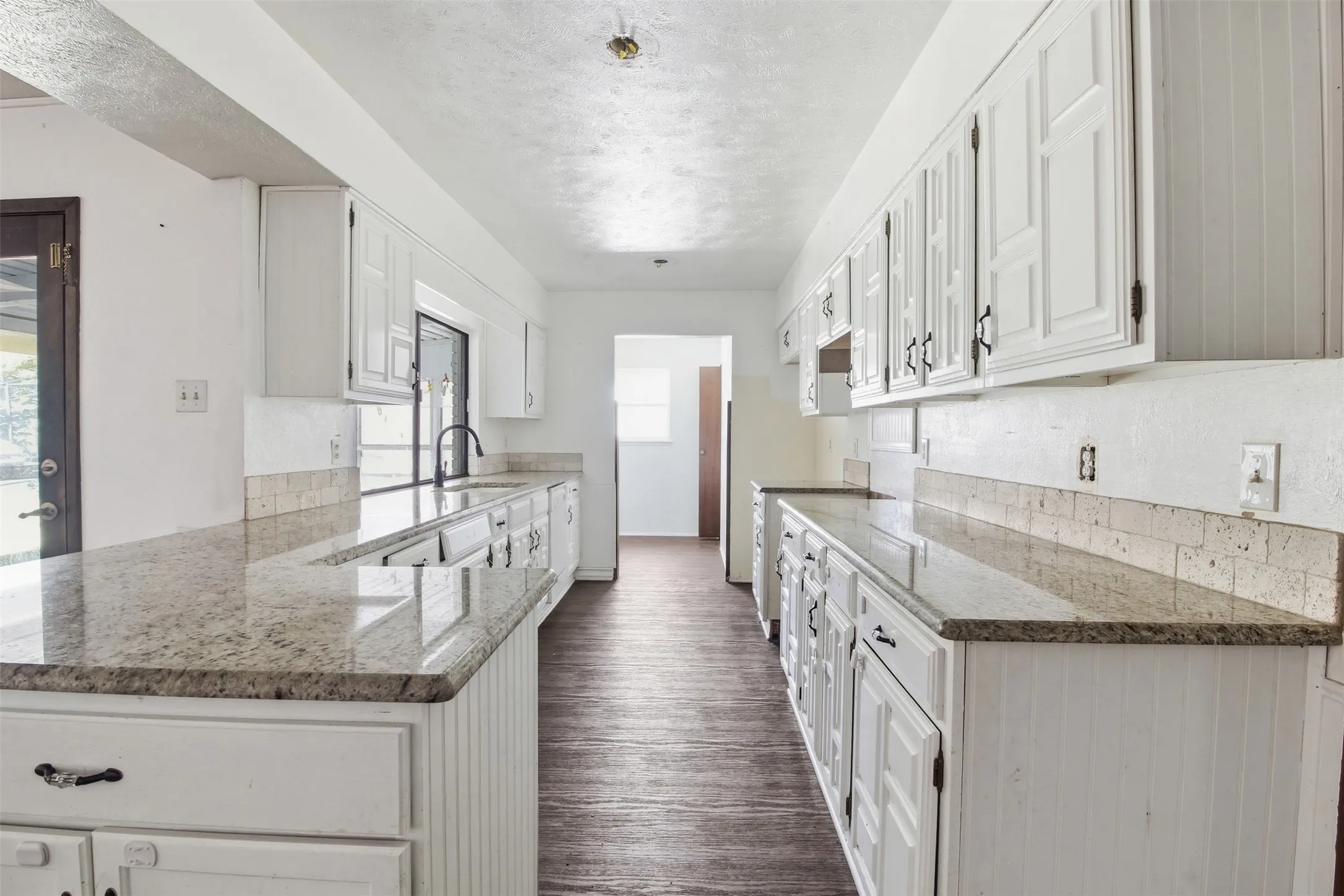 Galley kitchen featuring extensive granite countertops, white cabinetry, a built-in sink with a matte black faucet, a textured tile backsplash, and wood-finish flooring