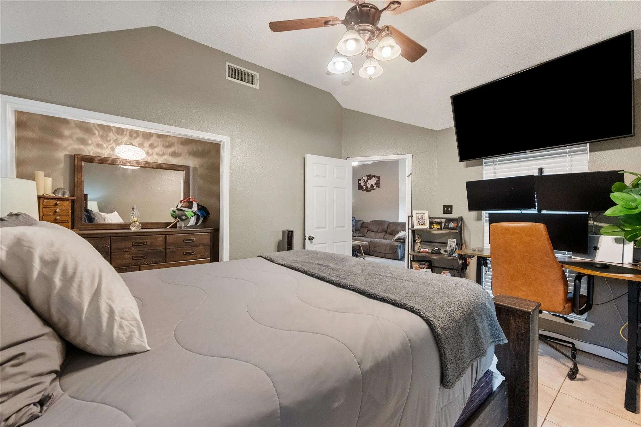 Vaulted ceiling bedroom featuring a ceiling fan with integrated lighting, a built-in wall niche with a light fixture, a desk area with a wall-mounted display, and an open doorway providing access to an additional room