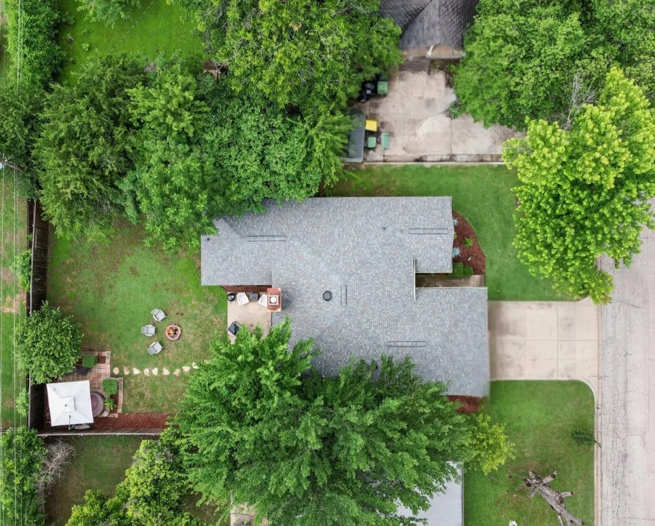 Overhead property view featuring a gabled roofline, expansive green lawn, mature perimeter trees, a rear patio with stone pavers, and a dedicated fire pit area