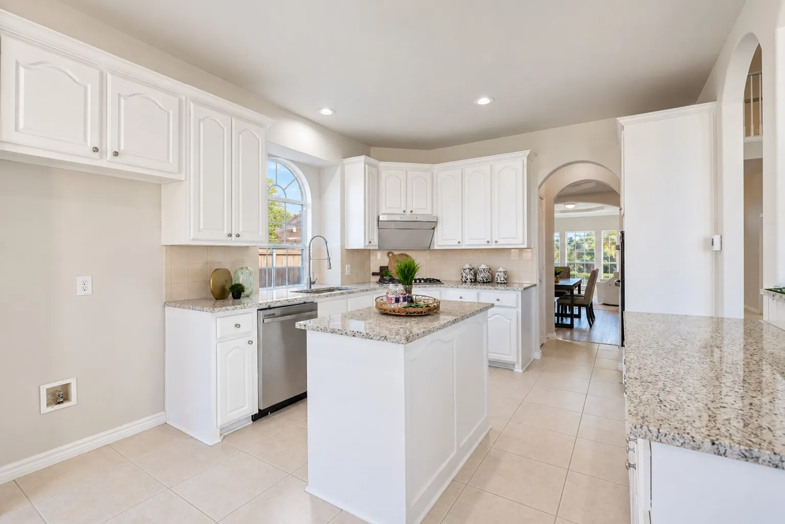 White cabinetry, stainless steel dishwasher, light-toned tile flooring, granite countertops, and an arched window