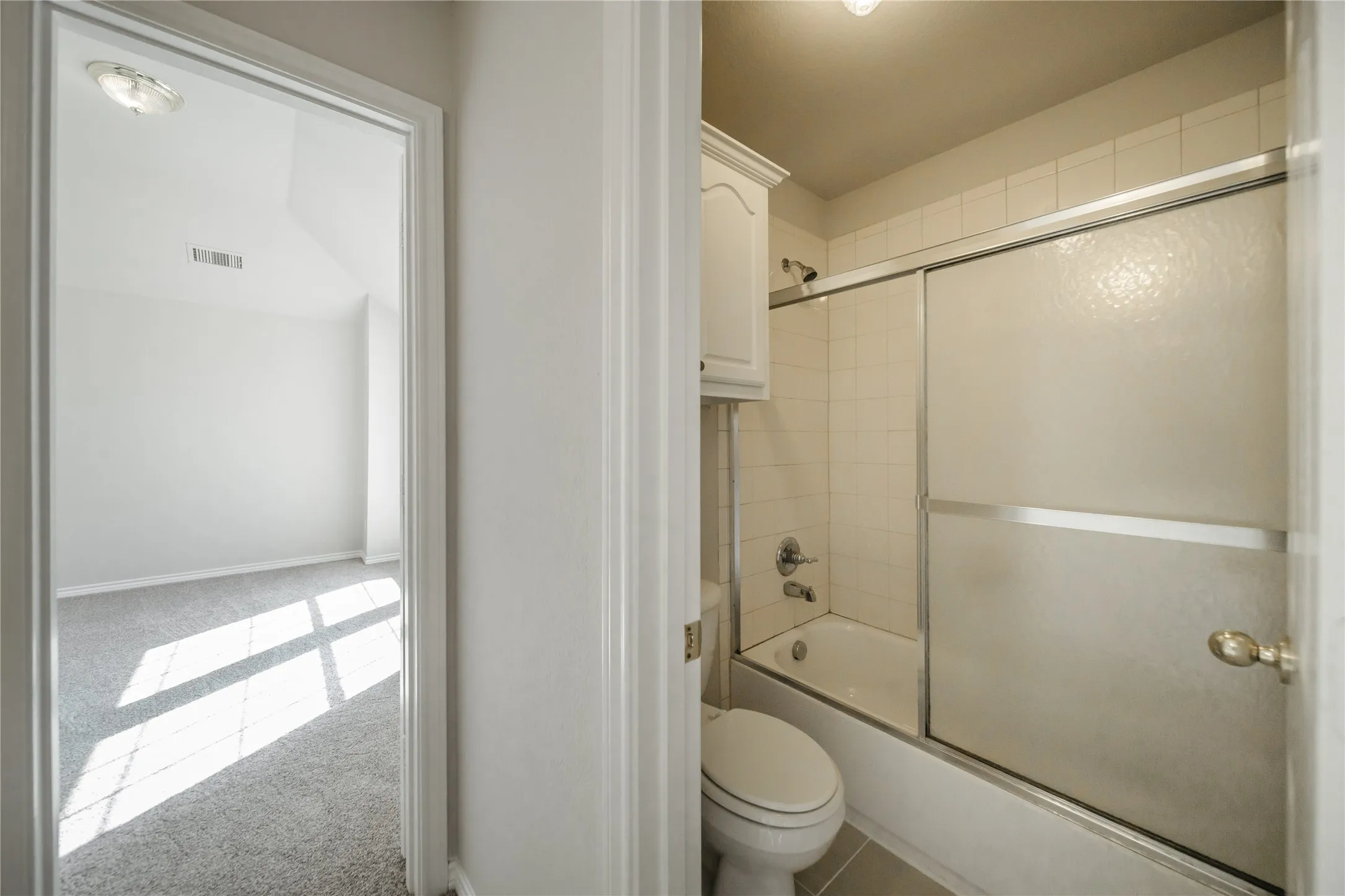 Bathroom featuring a shower-over-tub combination with sliding frosted glass doors, light-toned ceramic tile surround, and a built-in storage cabinet