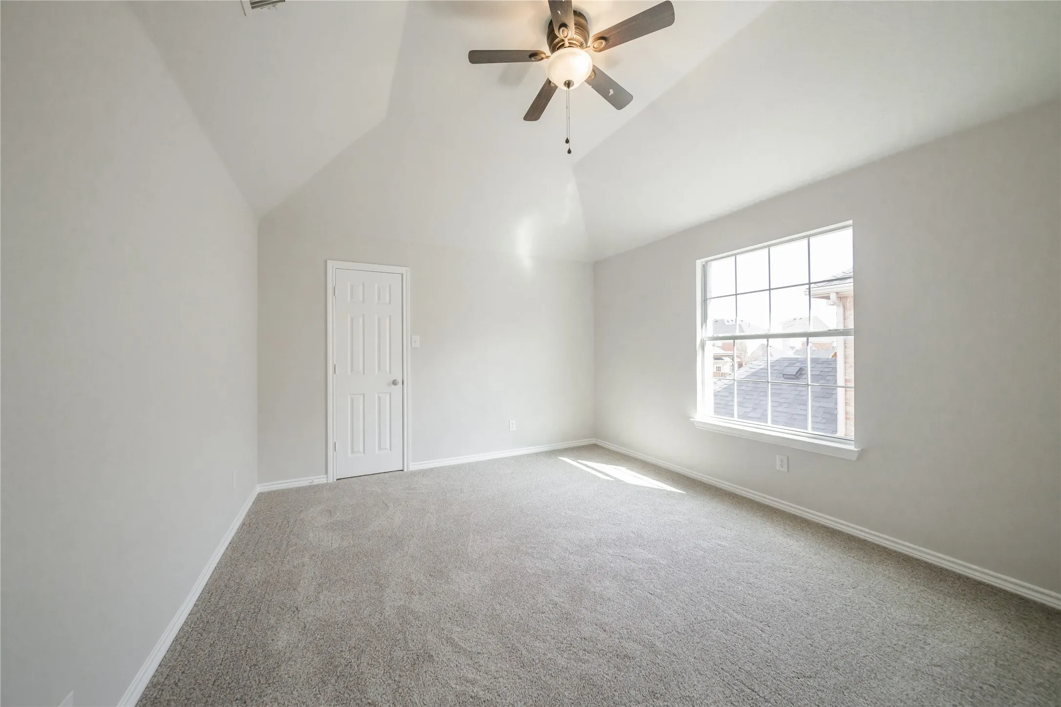 Spacious room featuring vaulted ceilings, a ceiling fan with integrated lighting, a large window with grid pattern, neutral carpeting, and a paneled interior door