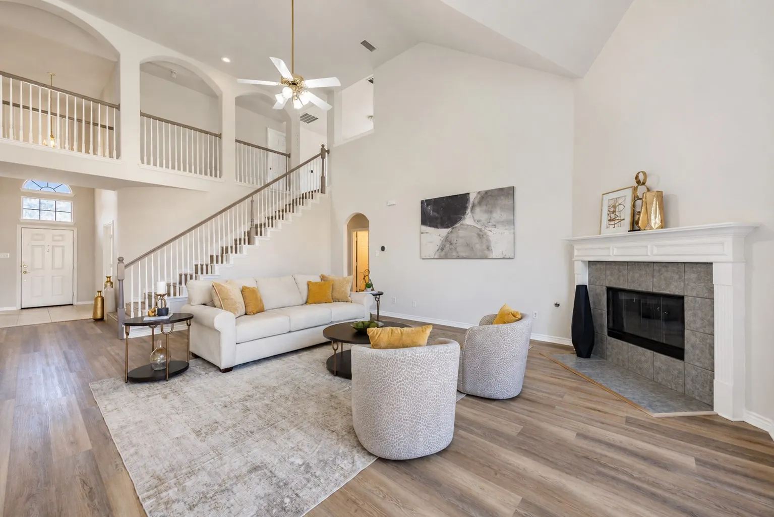 Vaulted ceiling with a ceiling fan, wood-finish flooring, two-story staircase with white balusters and wood handrail, fireplace with a tile surround and white mantel, and arched doorways