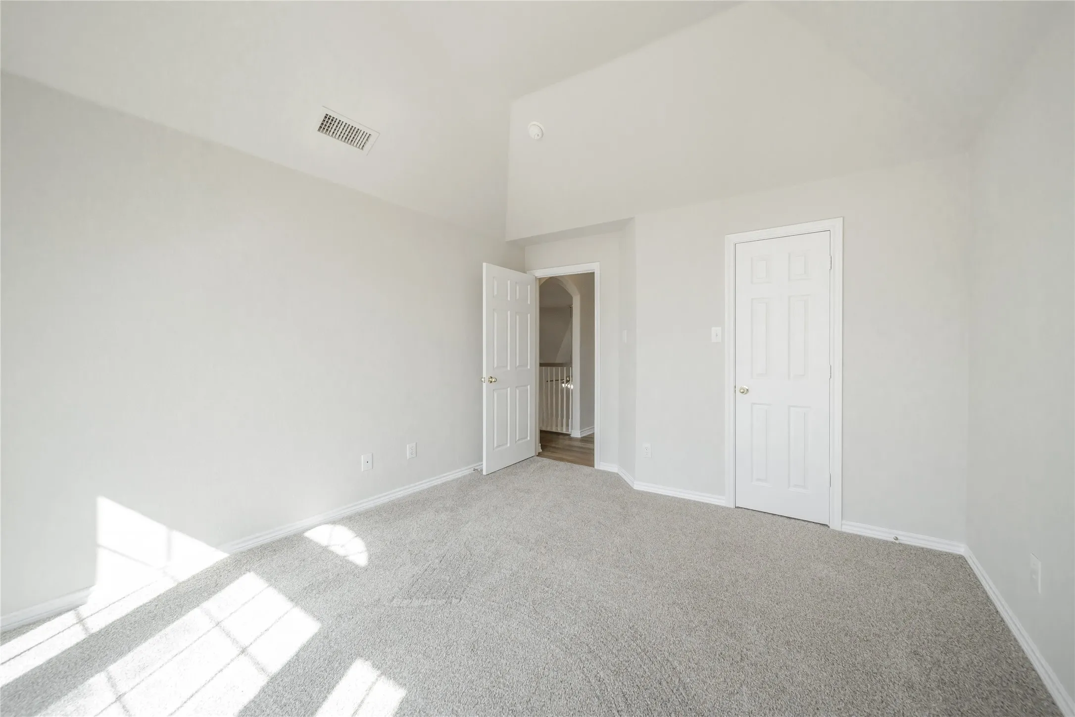 Spacious room featuring vaulted ceilings, light gray carpeting, fresh neutral paint, white trim, and a six-panel interior door