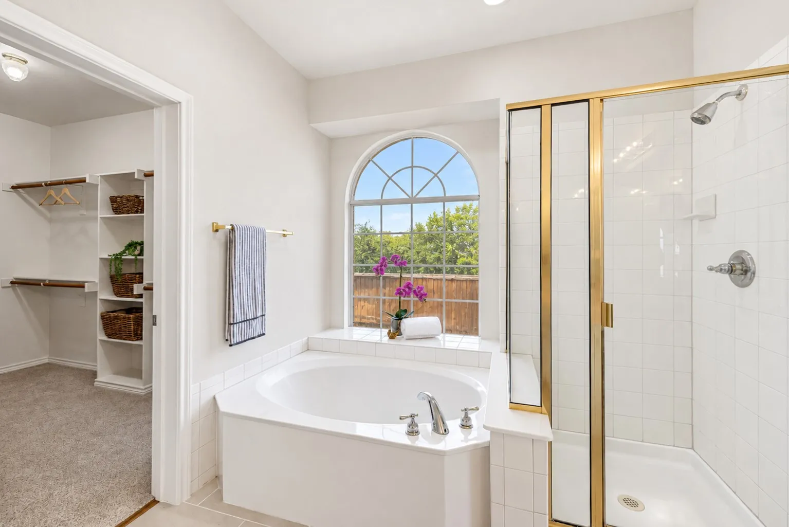 Soaking tub with an arched window, a glass-enclosed shower with gold-tone framing, and an adjacent walk-in closet with built-in shelving