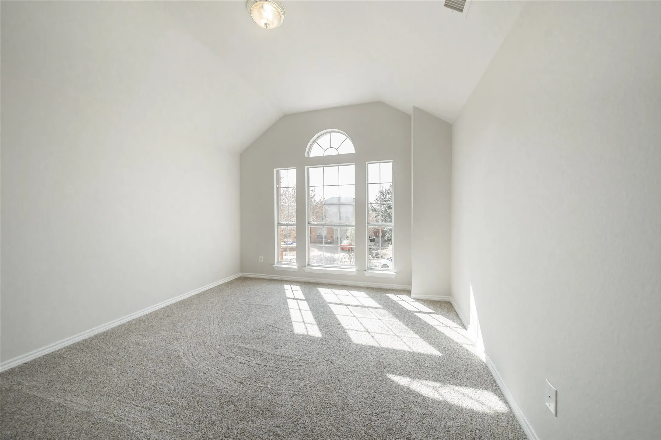 Arched picture window with transom detailing, vaulted ceiling, neutral gray carpet, white baseboards, and a flush mount ceiling light fixture