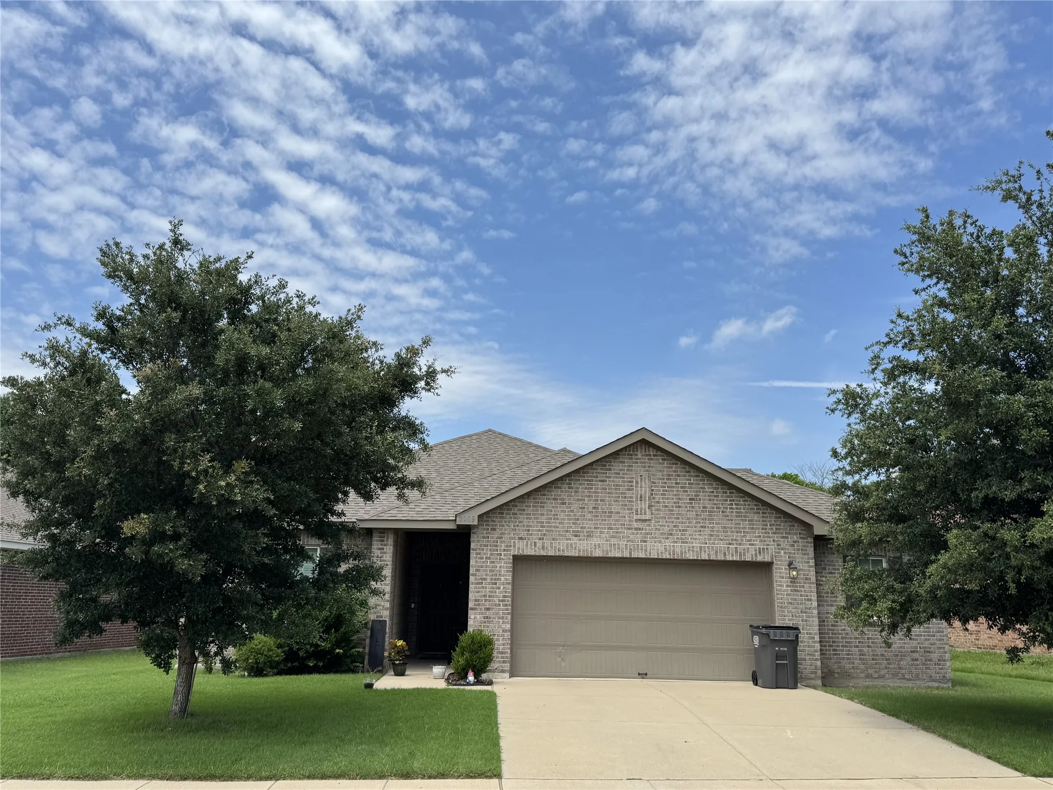 Brick exterior with a gabled roofline, front-facing garage, concrete driveway, and established trees