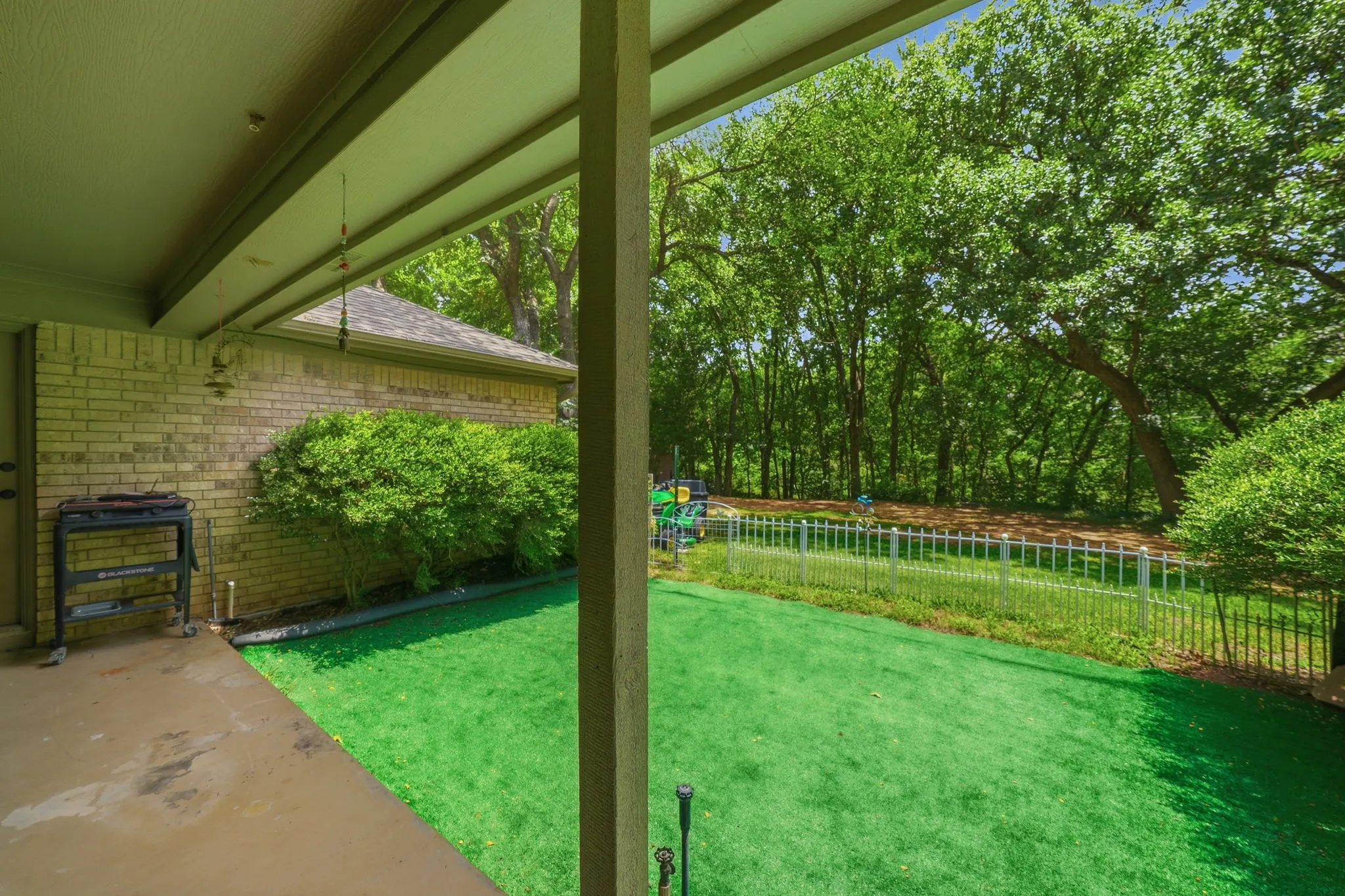 Covered patio with a concrete slab, overlooking a fenced yard featuring artificial turf and mature trees beyond