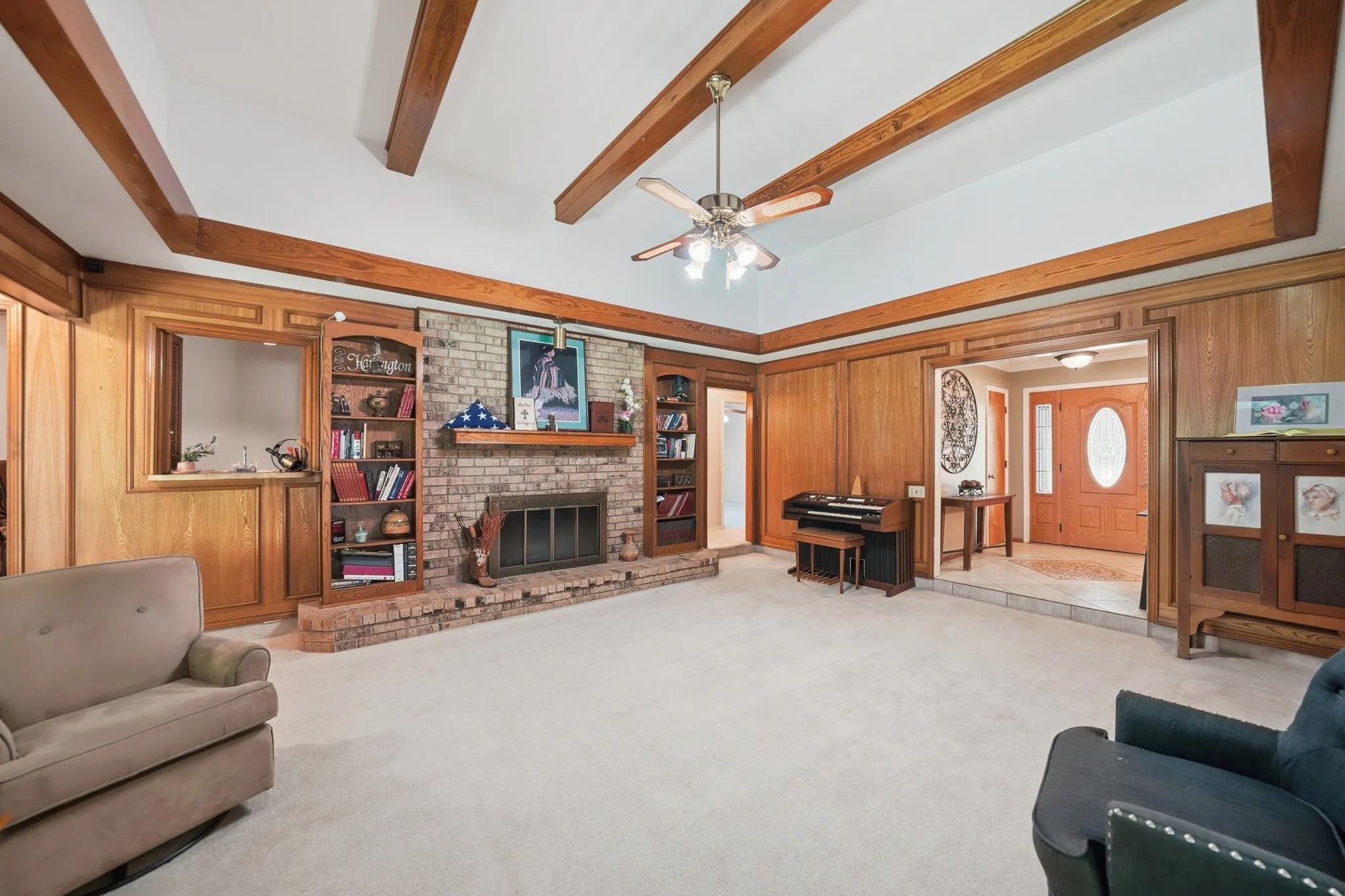 Spacious living area featuring a brick fireplace, wood paneling, exposed wood ceiling beams, and integrated shelving