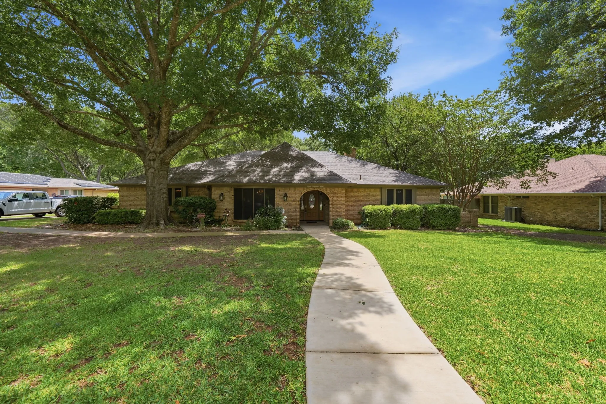 Brick exterior home featuring an arched entry, a concrete pathway, a large mature tree, and landscaped shrubbery