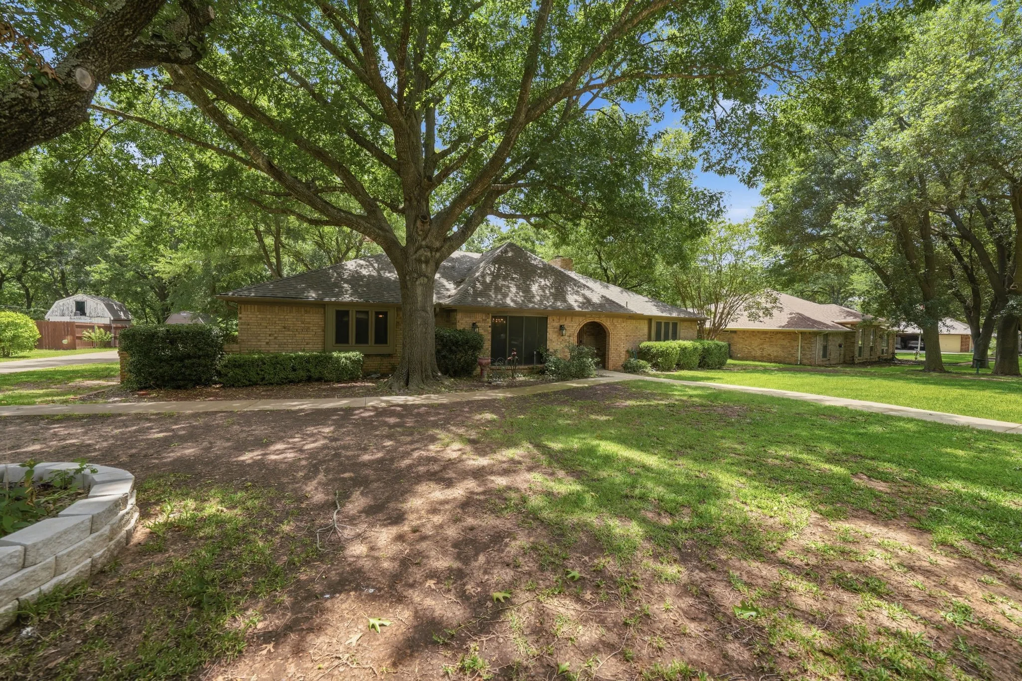 Brick exterior residence featuring a shaded front lawn, mature trees, a curved concrete pathway, and an additional outbuilding