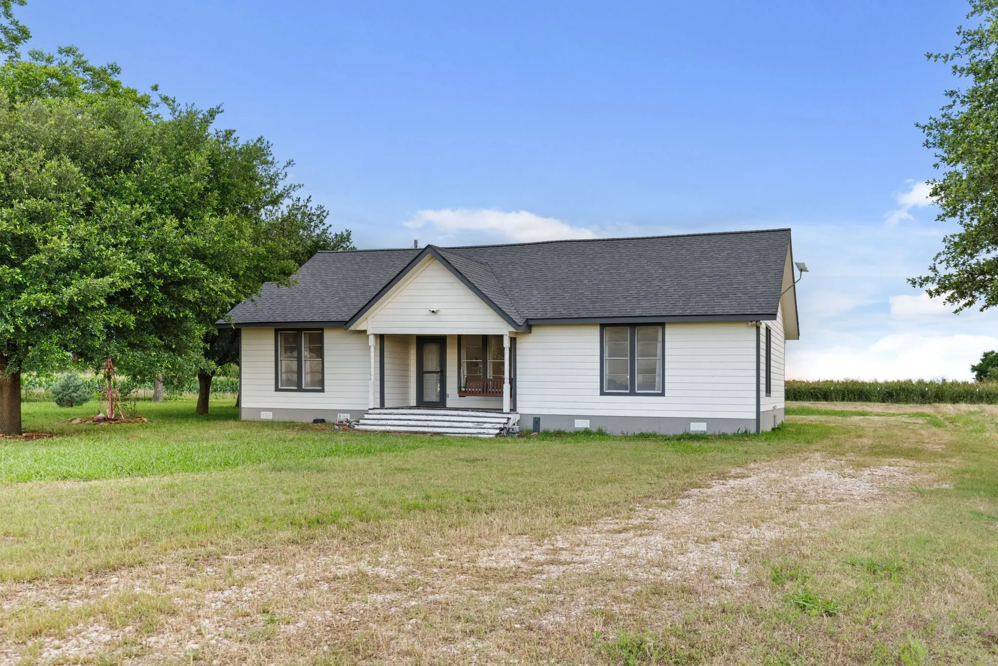 White siding exterior with dark trim windows, covered porch, dark shingle roof, and surrounding green space
