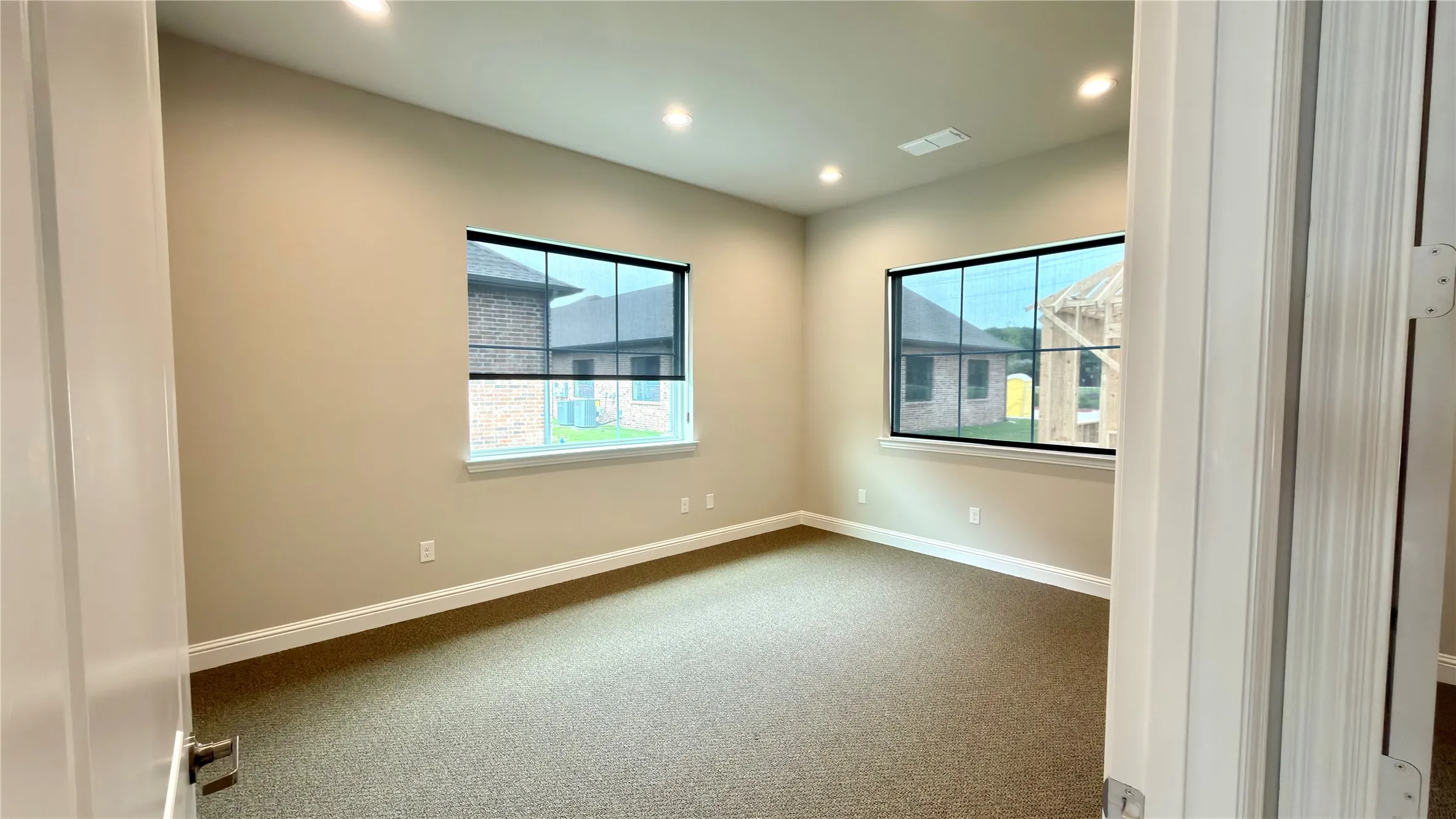 Neutral-toned room featuring a soft brown carpet, two large windows with roller shades, recessed ceiling lighting, and white baseboards