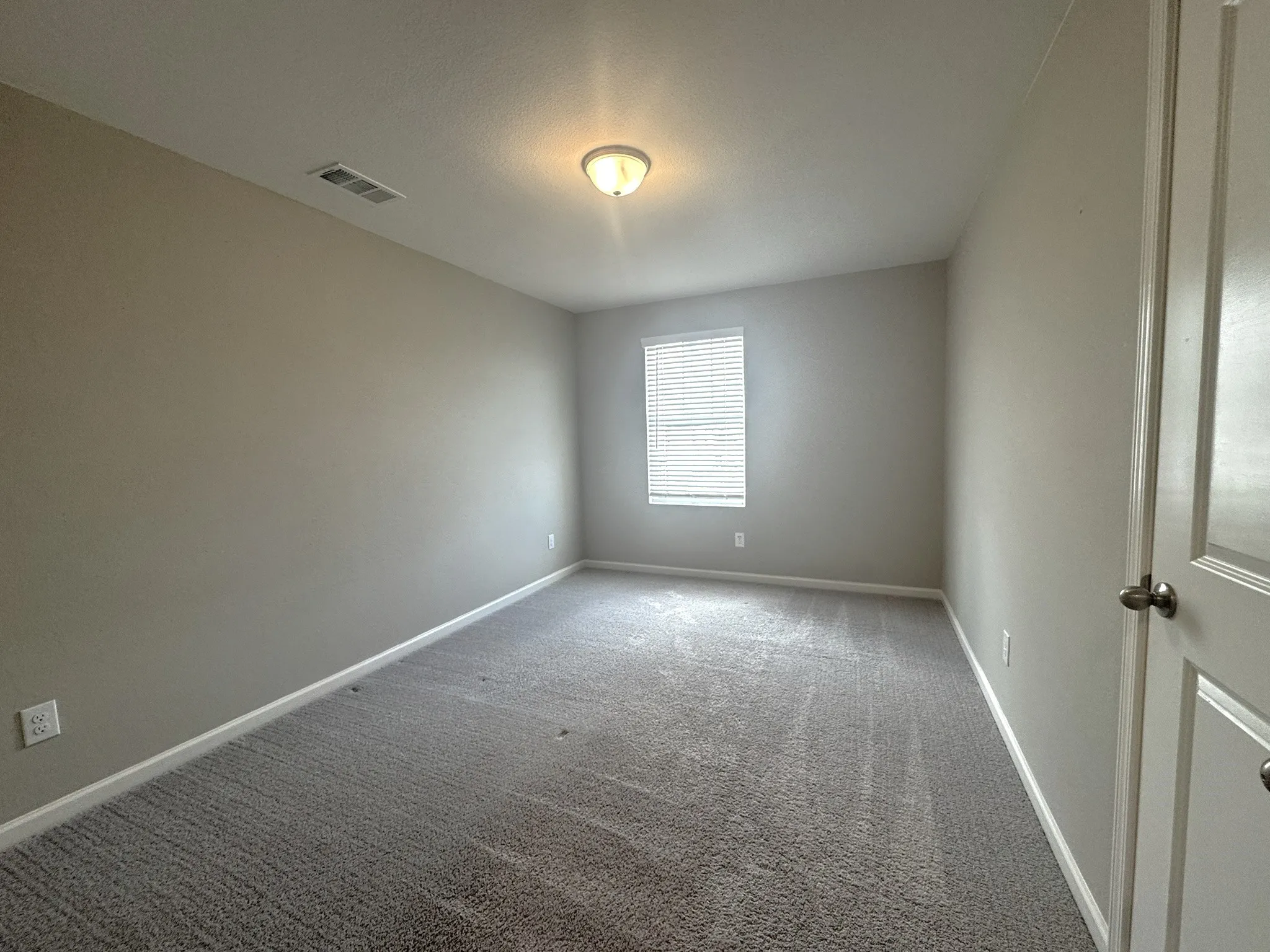 Carpeted room featuring neutral wall tones, white trim, a single window with blinds, a ceiling-mounted light fixture, and a white interior door with a silver knob