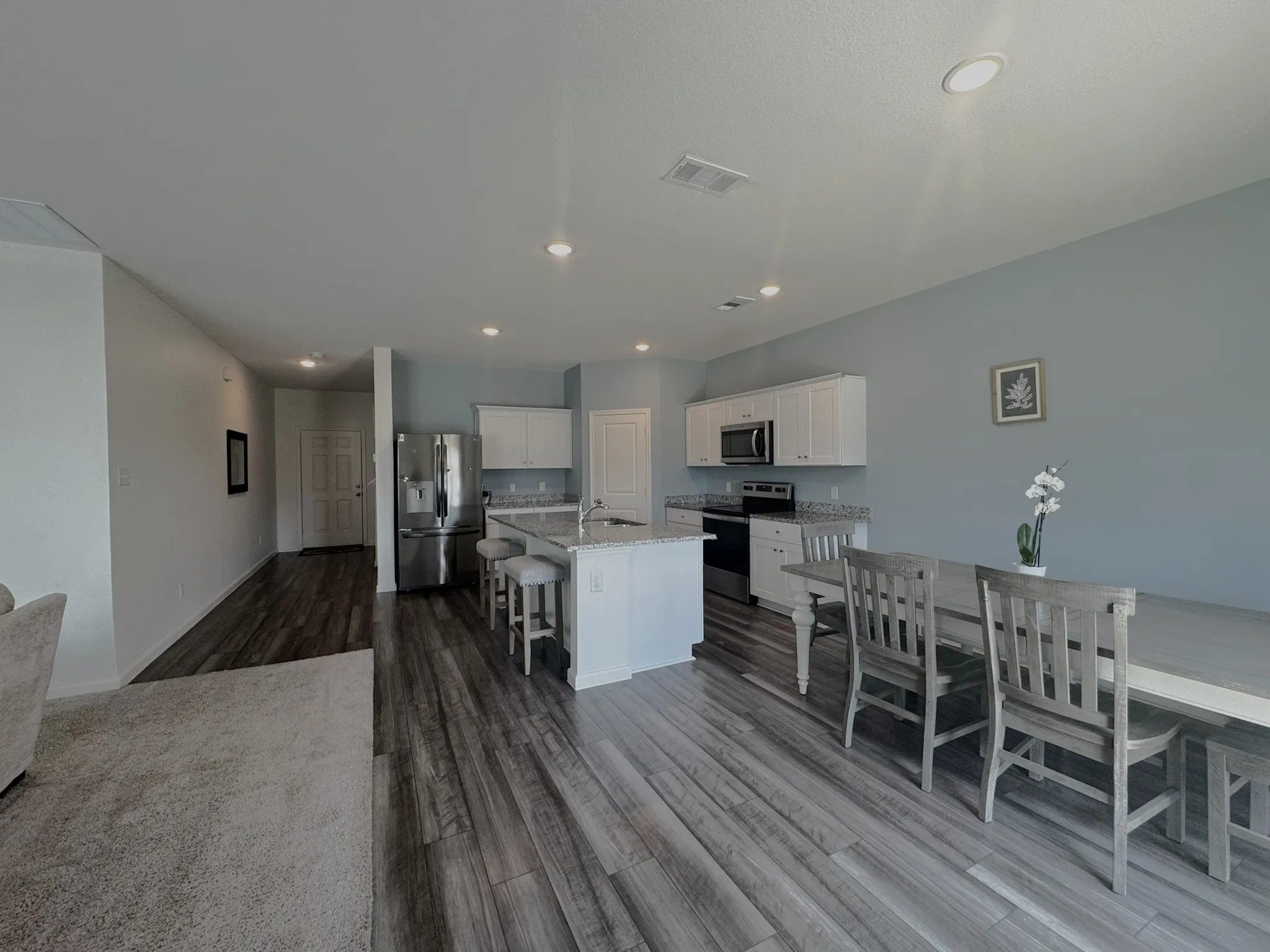 Open-concept kitchen featuring wood-finish flooring, white cabinetry, granite-style countertops with a matching backsplash, stainless steel appliances, and an island with bar seating