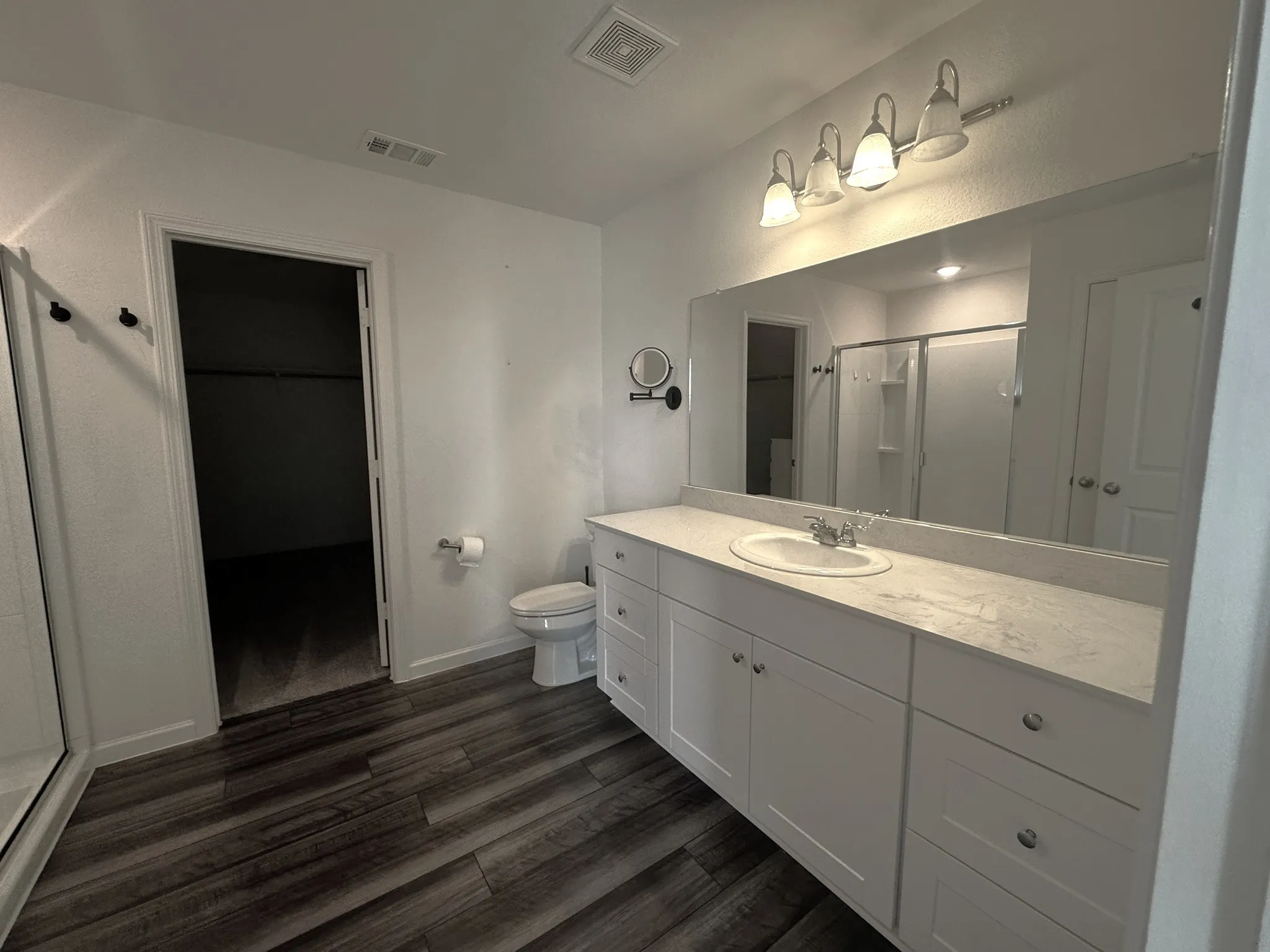 Spacious bathroom featuring a double vanity with white cabinetry and a light-toned countertop, wood-finish flooring, a toilet, a walk-in shower with clear glass enclosure, and an open closet space