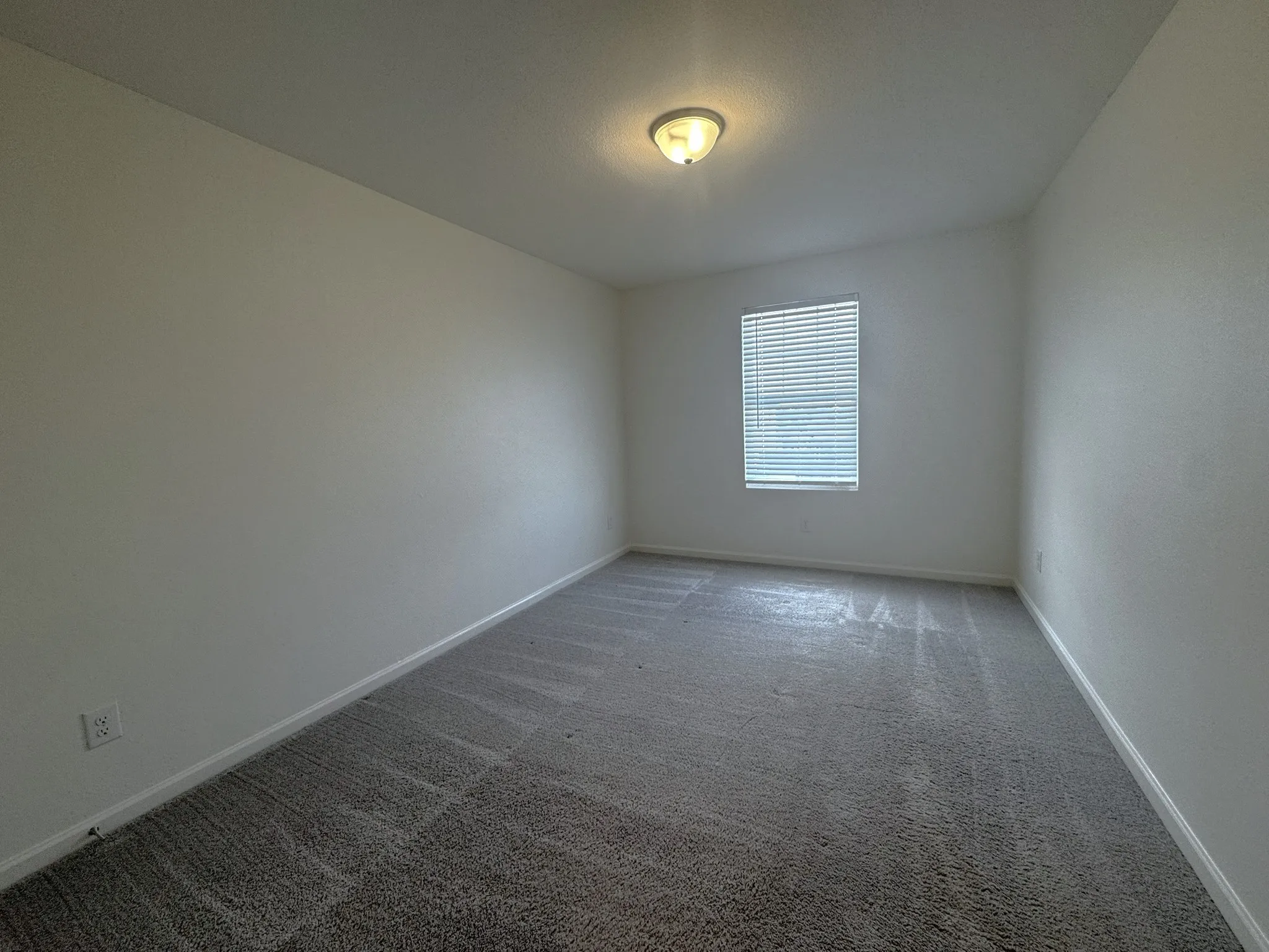 Neutral-toned room featuring a single window with horizontal blinds, overhead flush mount lighting, and gray patterned carpeting