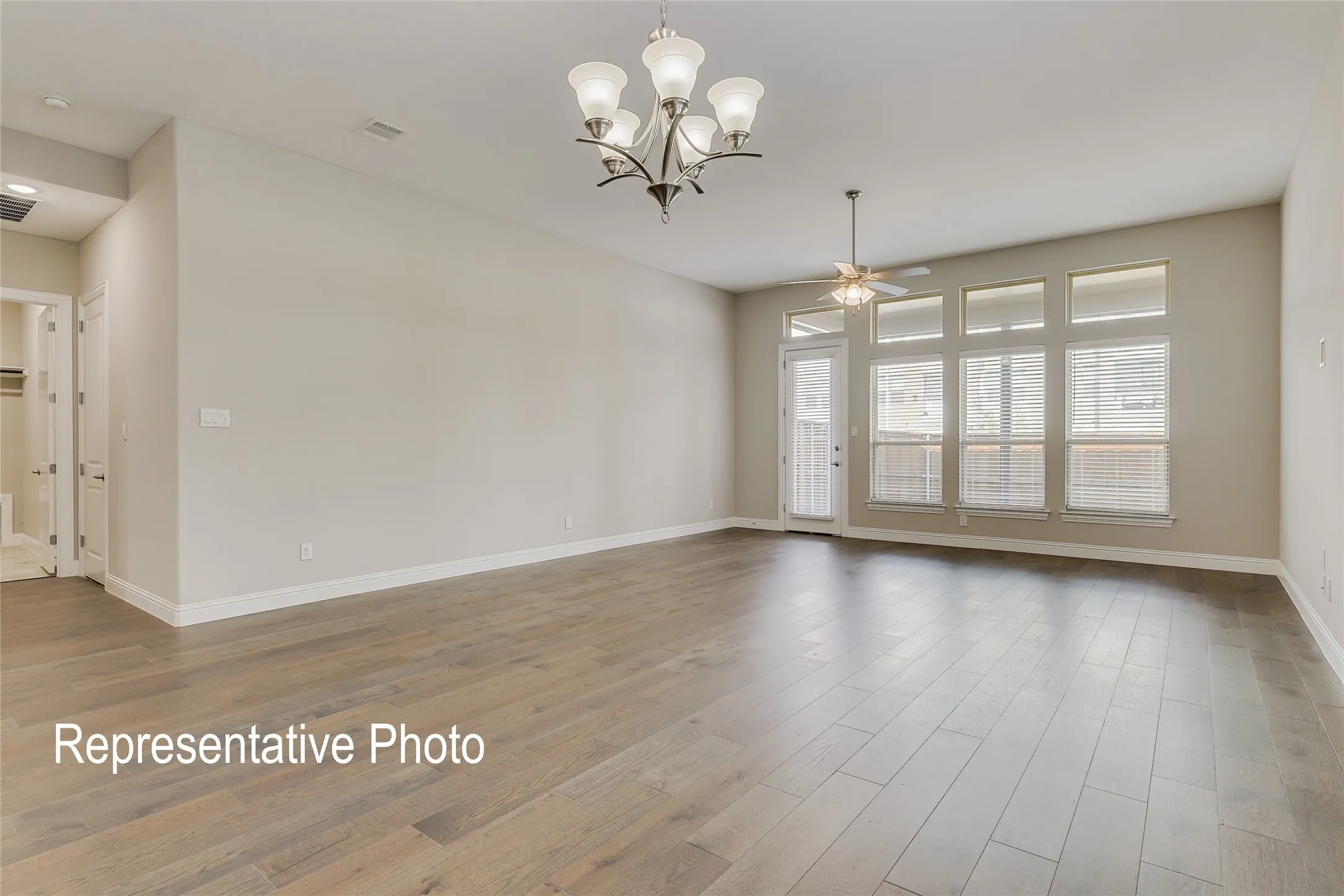 Spacious room featuring wood-finish flooring, a contemporary chandelier, a ceiling fan, multiple windows with blinds, and a white trim baseboard
