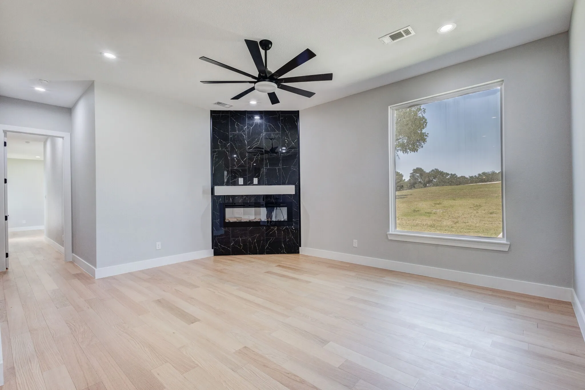 Spacious room featuring light wood-finish flooring, a contemporary black marbled fireplace surround with a white mantel, and a multi-blade ceiling fan