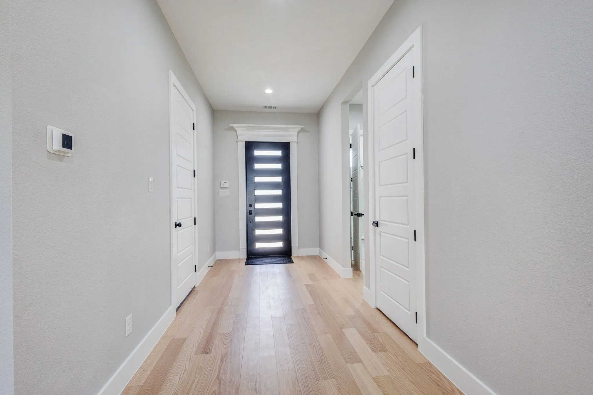 Entrance hallway featuring wood-finish flooring, a contemporary black entry door with frosted glass panels, white interior doors with black hardware, recessed ceiling lighting, and crisp white baseboards