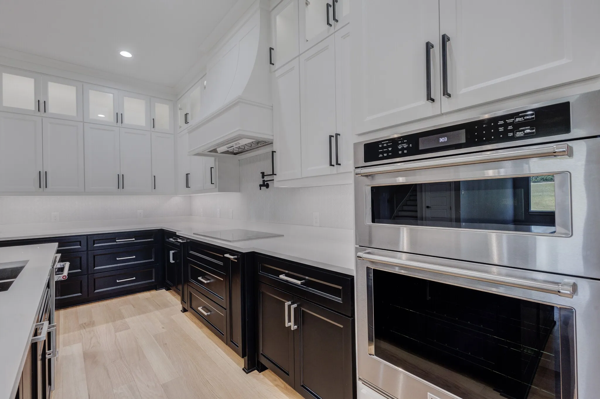 Gourmet kitchen featuring a built-in stainless steel double oven, sleek cooktop, contrasting black and white cabinetry, recessed lighting, and wood-finish flooring