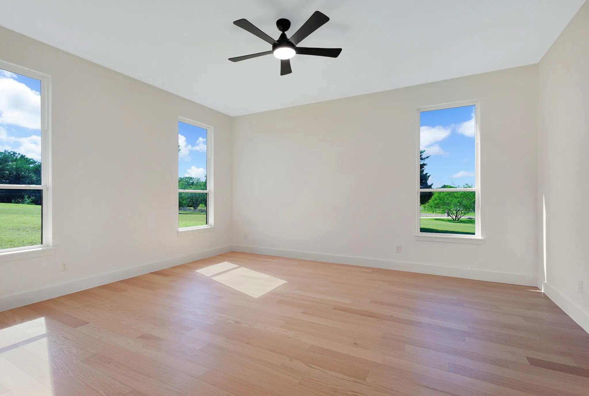 Spacious room featuring light wood-finish flooring, three vertical windows, white baseboards, and a black ceiling fan with integrated lighting
