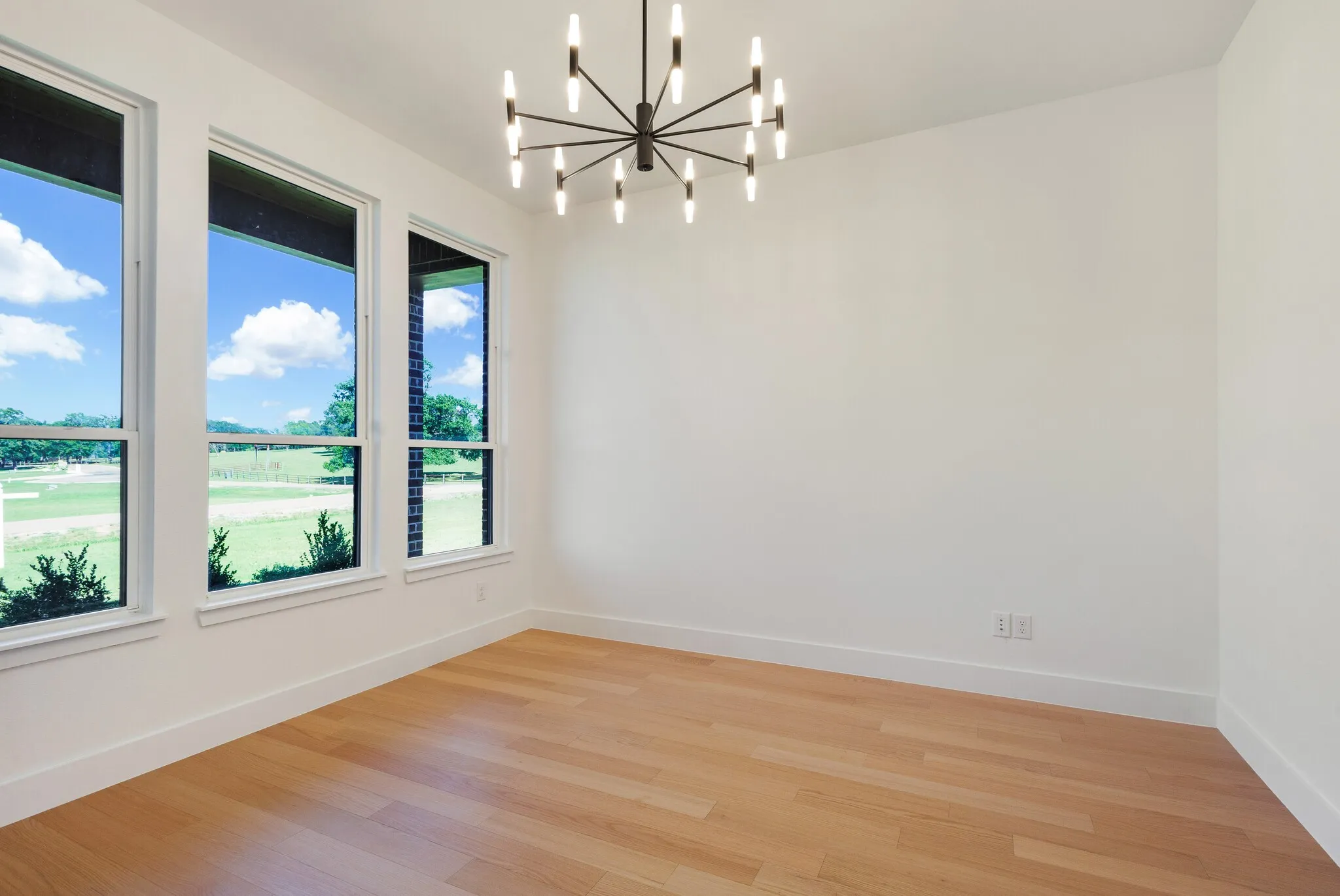 Spacious room featuring three large windows, a contemporary chandelier, light wood-finish flooring, and crisp white baseboards