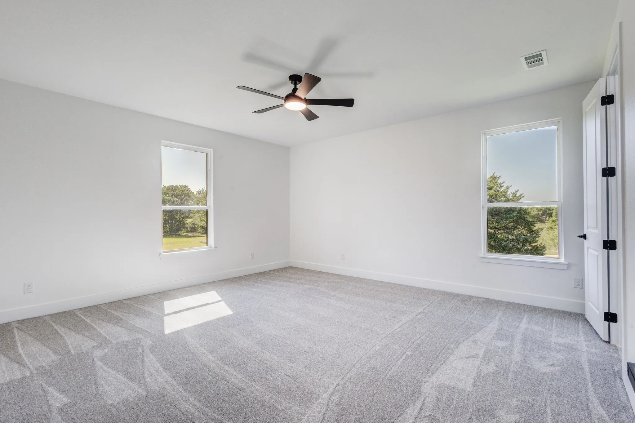 Bright room with white painted walls, gray patterned carpet, a ceiling fan with integrated lighting, and two single-pane windows