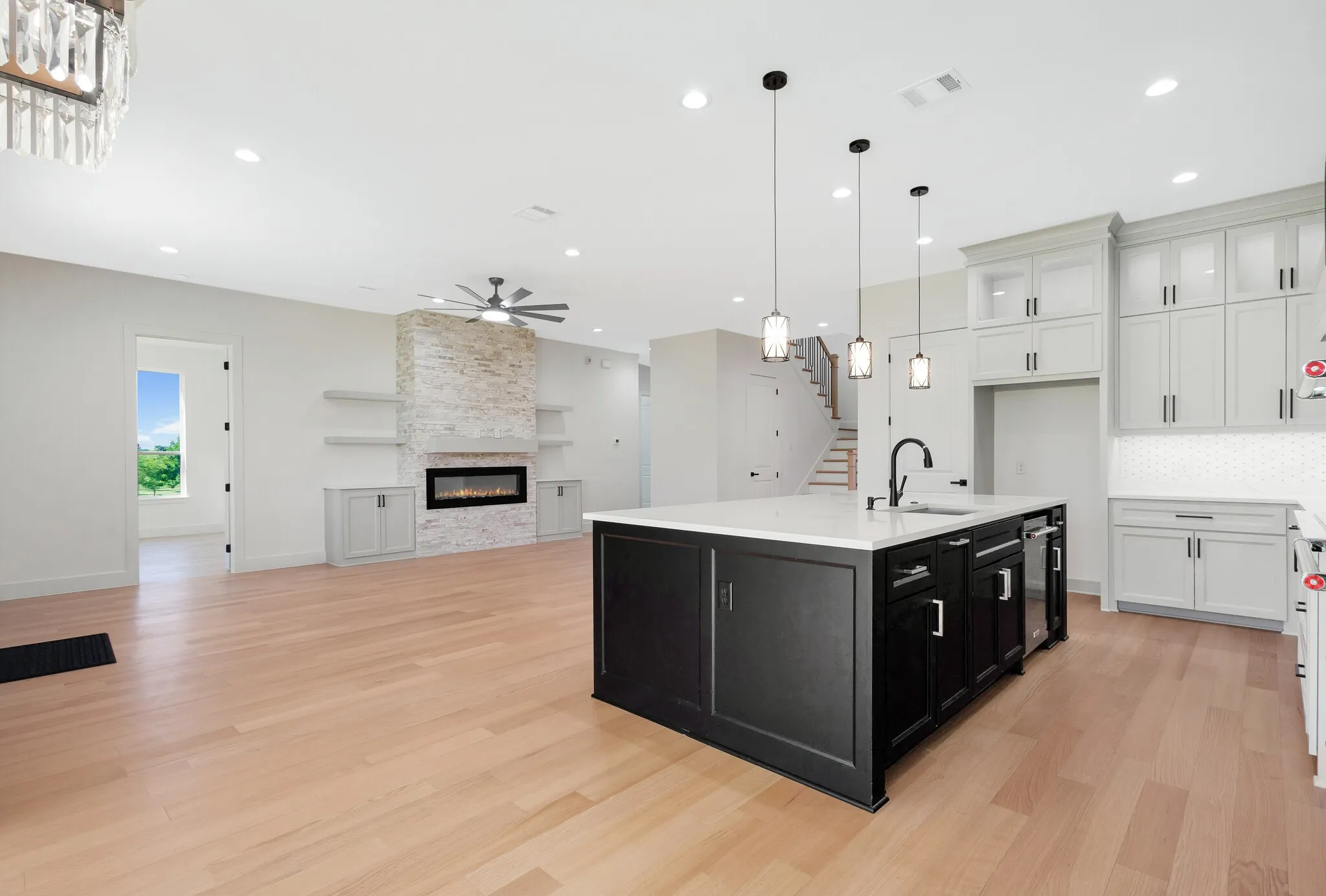 Spacious kitchen and living area featuring wood-finish flooring, a central kitchen island with a sink and pendant lighting, white shaker cabinetry with tile backsplash, a stacked stone fireplace with built-in shelving and cabinetry, and recessed lighting throughout
