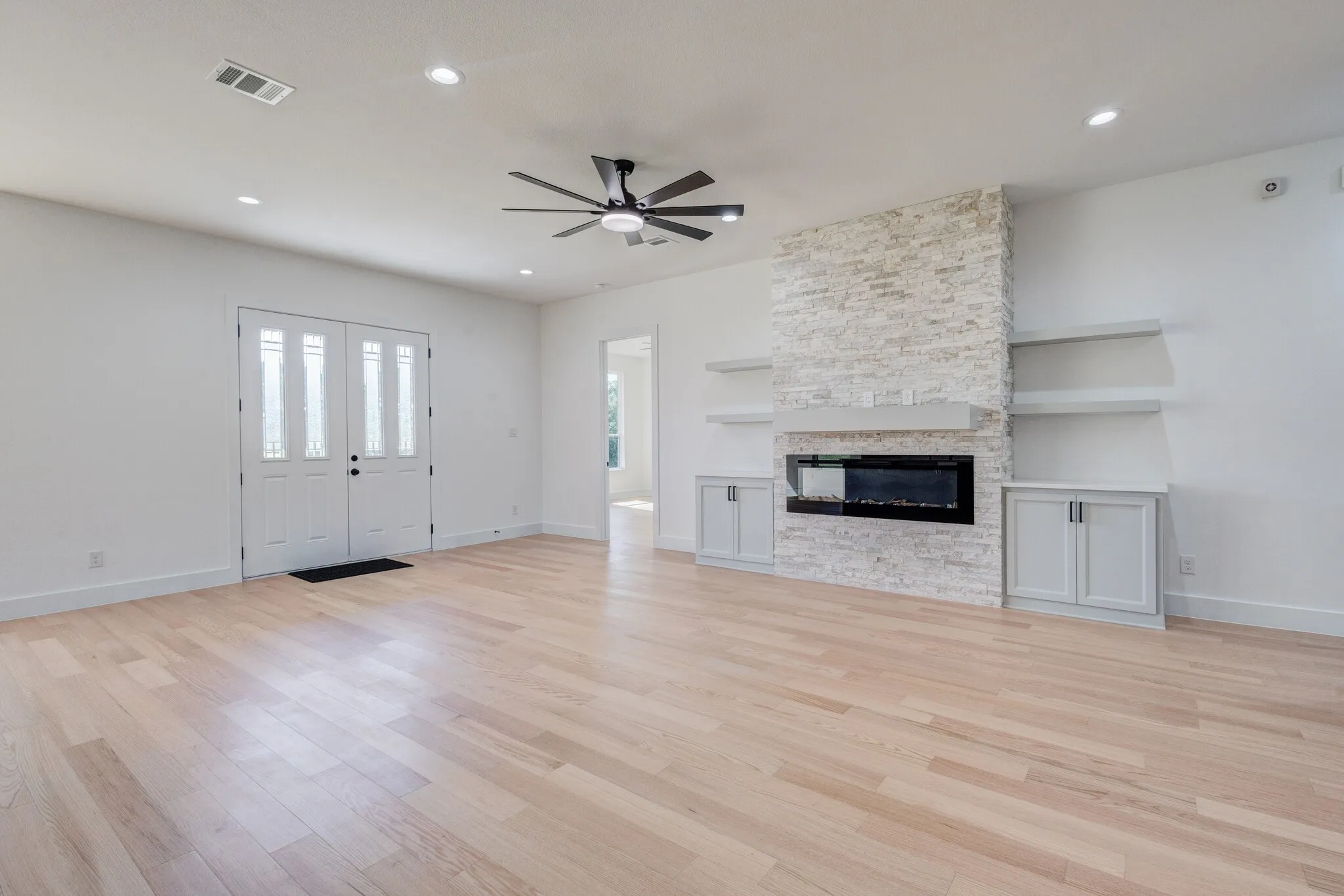 Spacious living area featuring light wood-finish flooring, a stone-clad fireplace with an electric insert, built-in cabinetry, floating shelves, and recessed lighting
