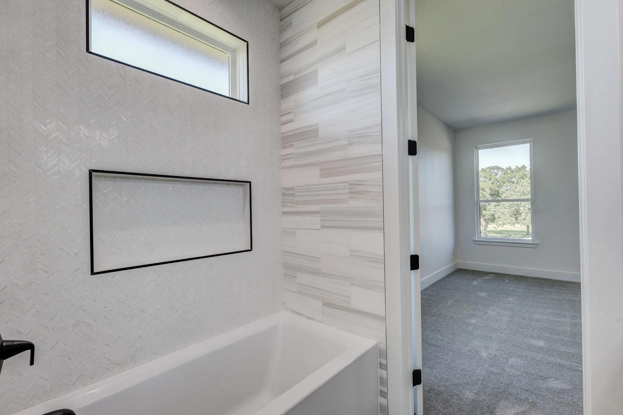 White soaking tub with textured white tile surround, black-framed shower niche, and rectangular transom window
