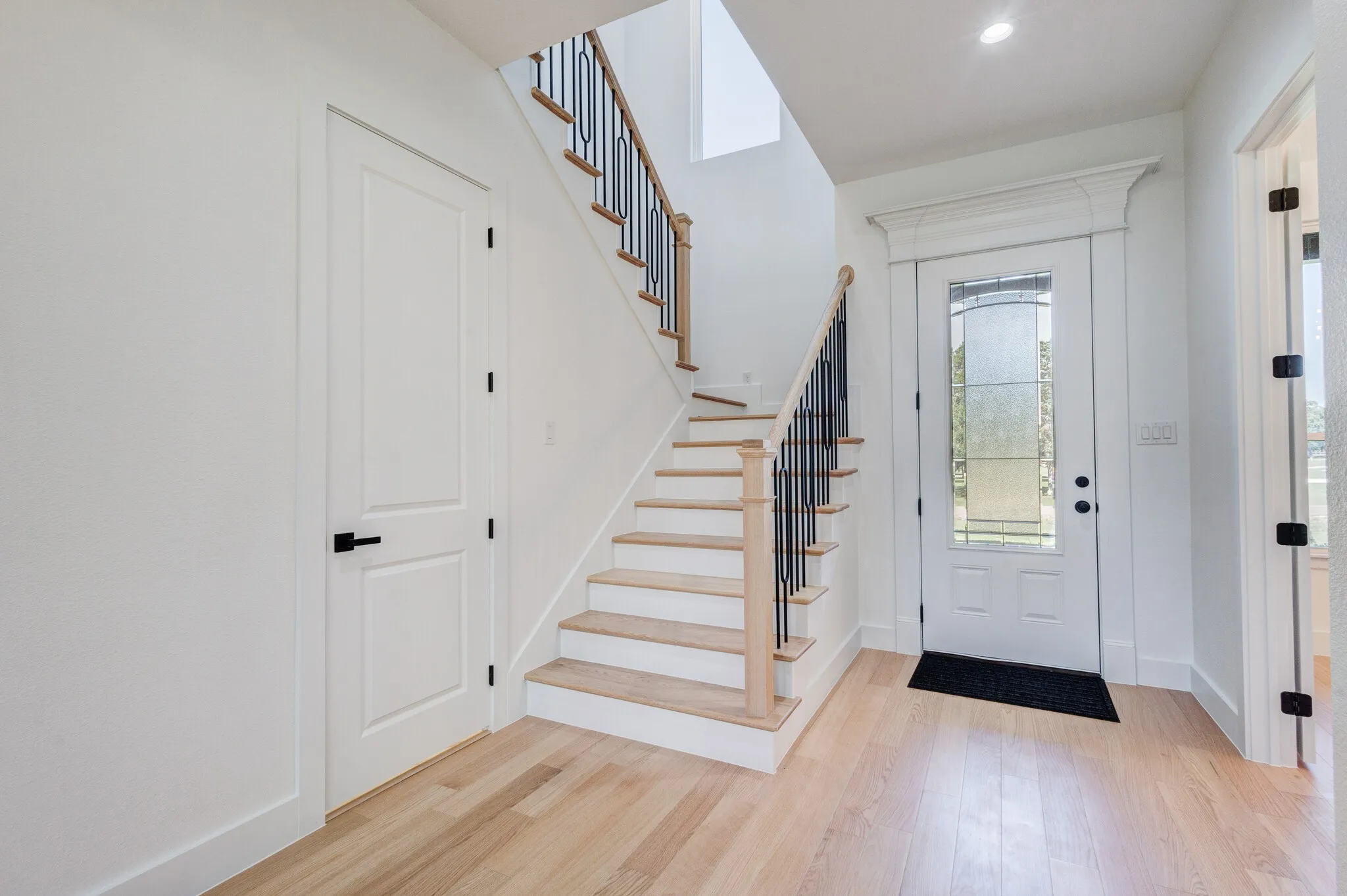 Entryway featuring light wood-finish flooring, a staircase with black metal balusters and wood-finish handrail, white paneled doors with black hardware, a recessed ceiling light, and a white front door with decorative glass inserts