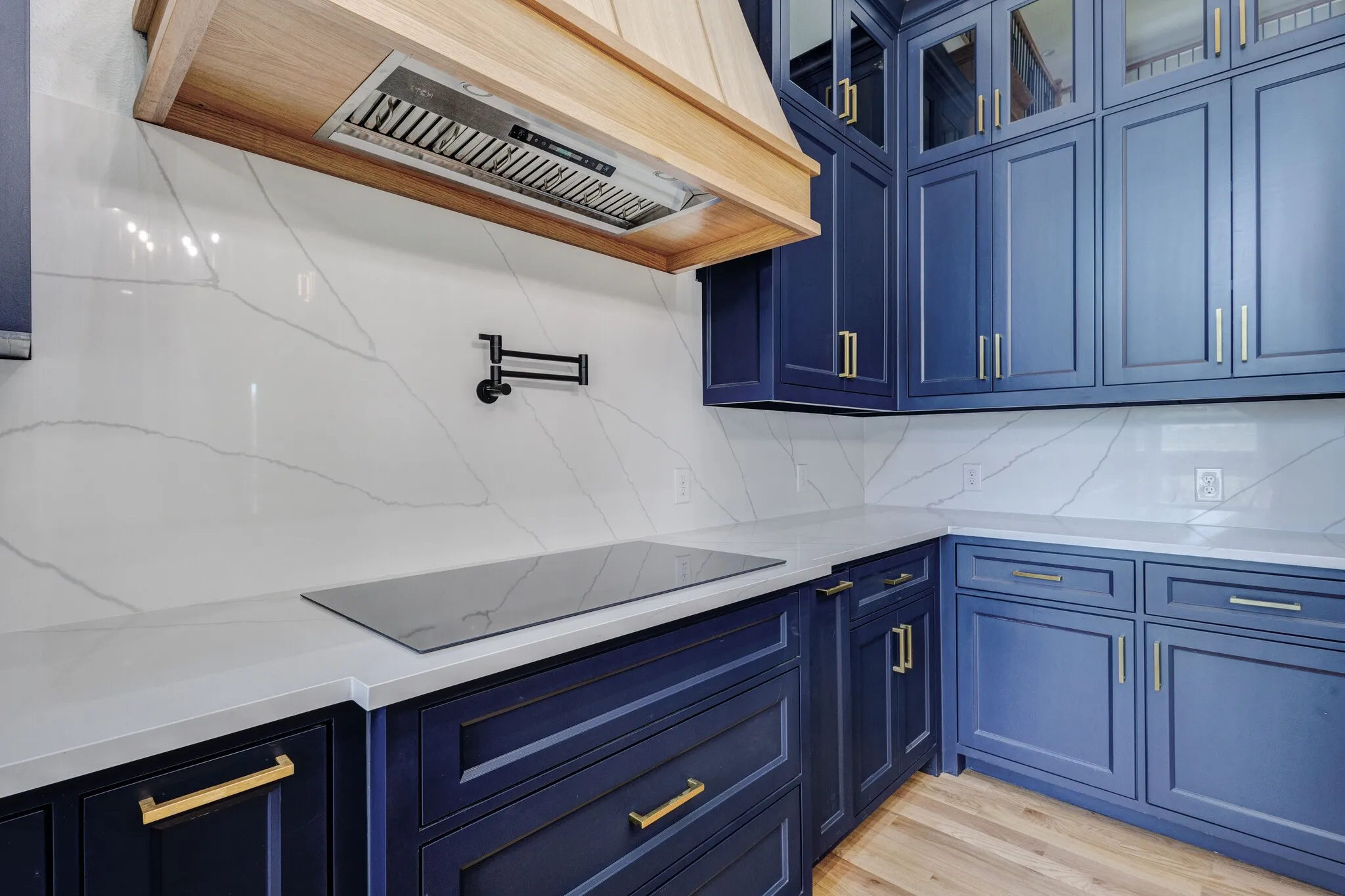 Kitchen featuring custom blue cabinetry with gold-tone hardware, a wood-finish range hood, white countertops, a contrasting white backsplash, and an integrated cooktop