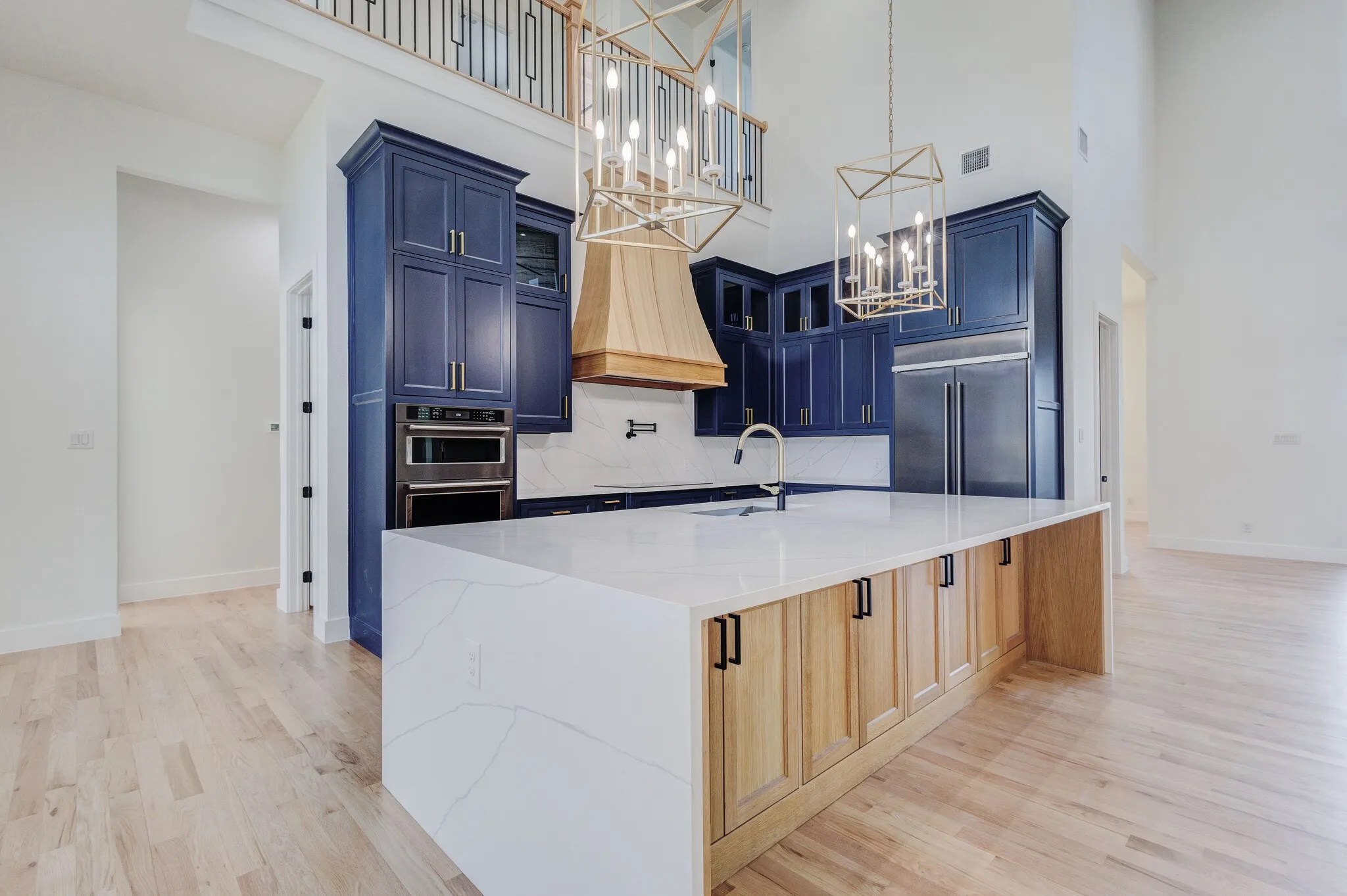 Spacious kitchen featuring a large central island with a waterfall countertop, two-tone cabinetry in blue and natural wood, stainless steel built-in appliances, light wood-finish flooring, and high ceilings with decorative light fixtures
