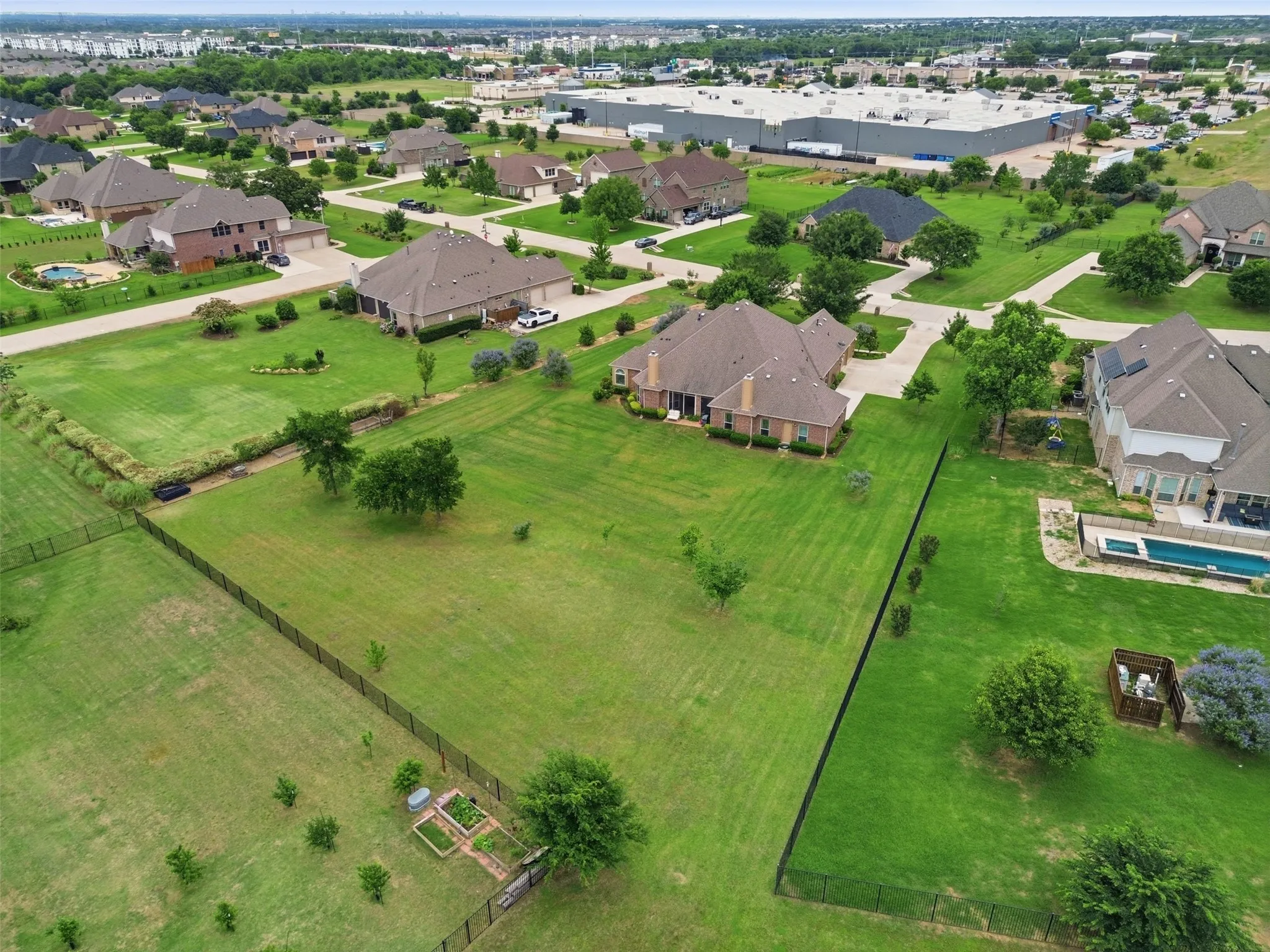 Expansive green lot featuring a black metal fence, mature trees, and an established garden plot