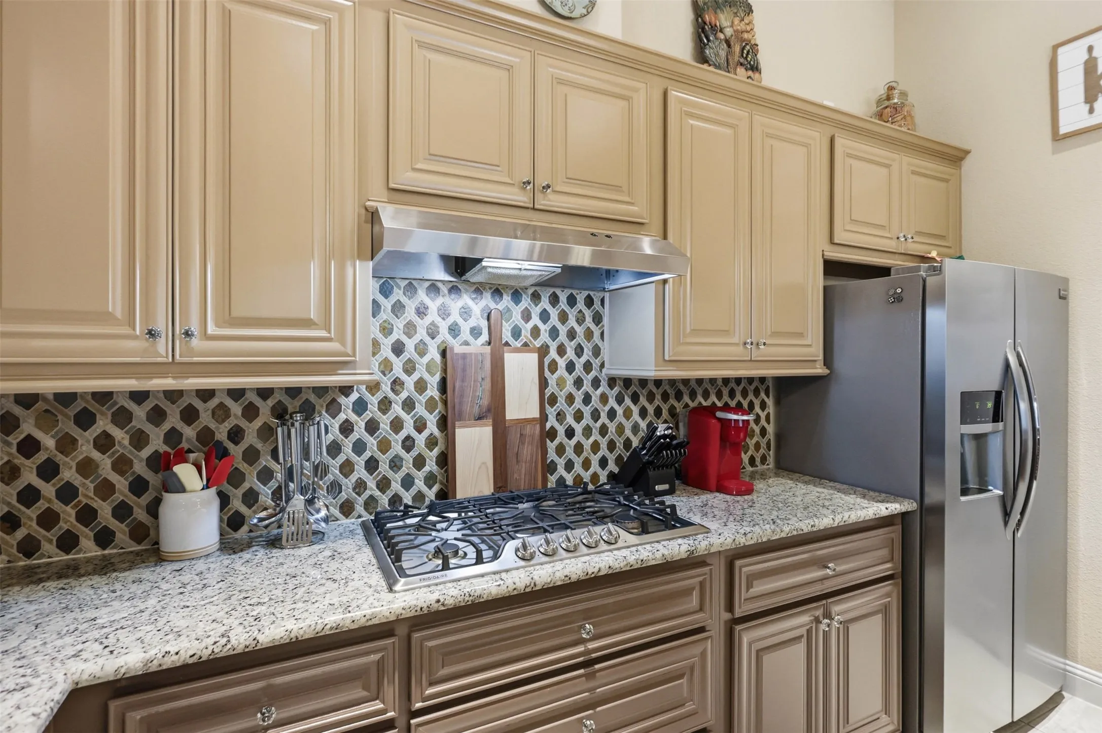 Kitchen featuring a gas range, stainless steel range hood, stone mosaic tile backsplash, granite countertops, and two-tone cabinetry