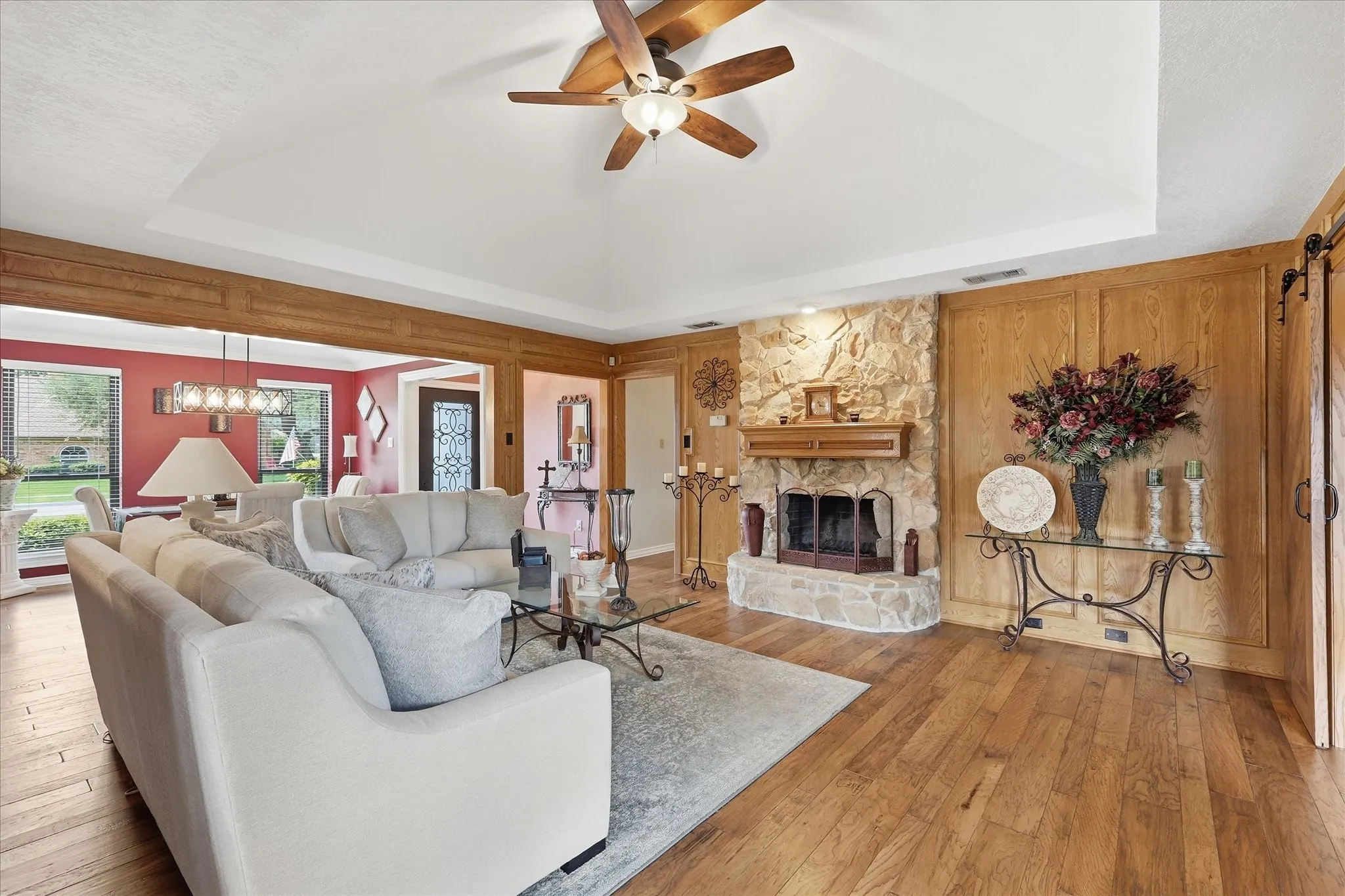Living room featuring a stone fireplace with wood mantel, wood-finish flooring, and a tray ceiling with a ceiling fan