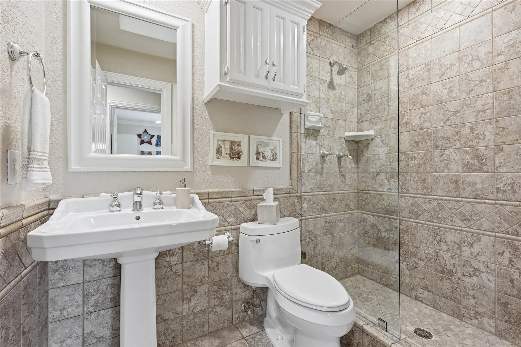 Pedestal sink with chrome fixtures, framed mirror, and white overhead cabinetry