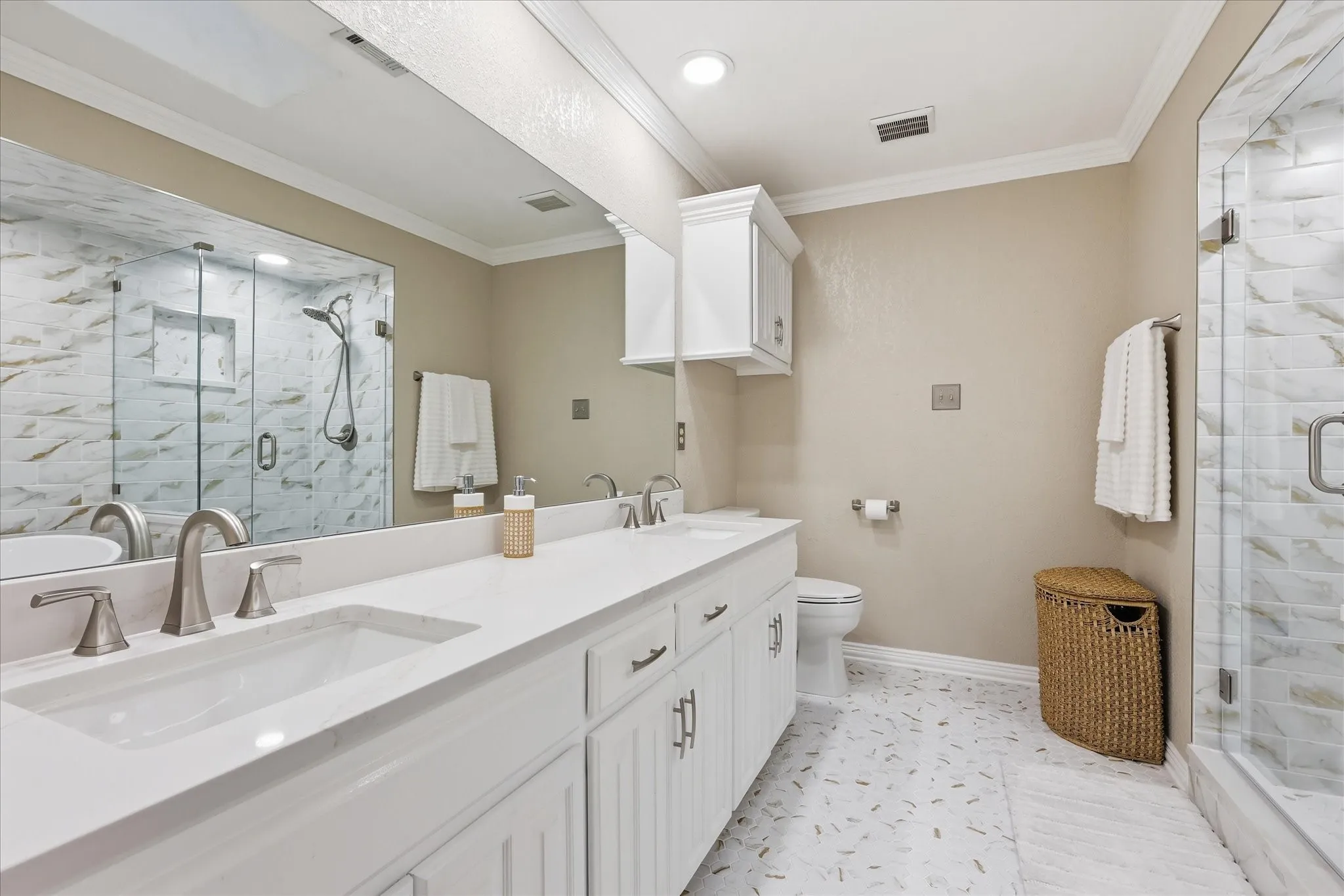 Bathroom featuring a dual vanity with white cabinetry and integrated sinks, a glass-enclosed shower with tiled walls, and a patterned tile floor