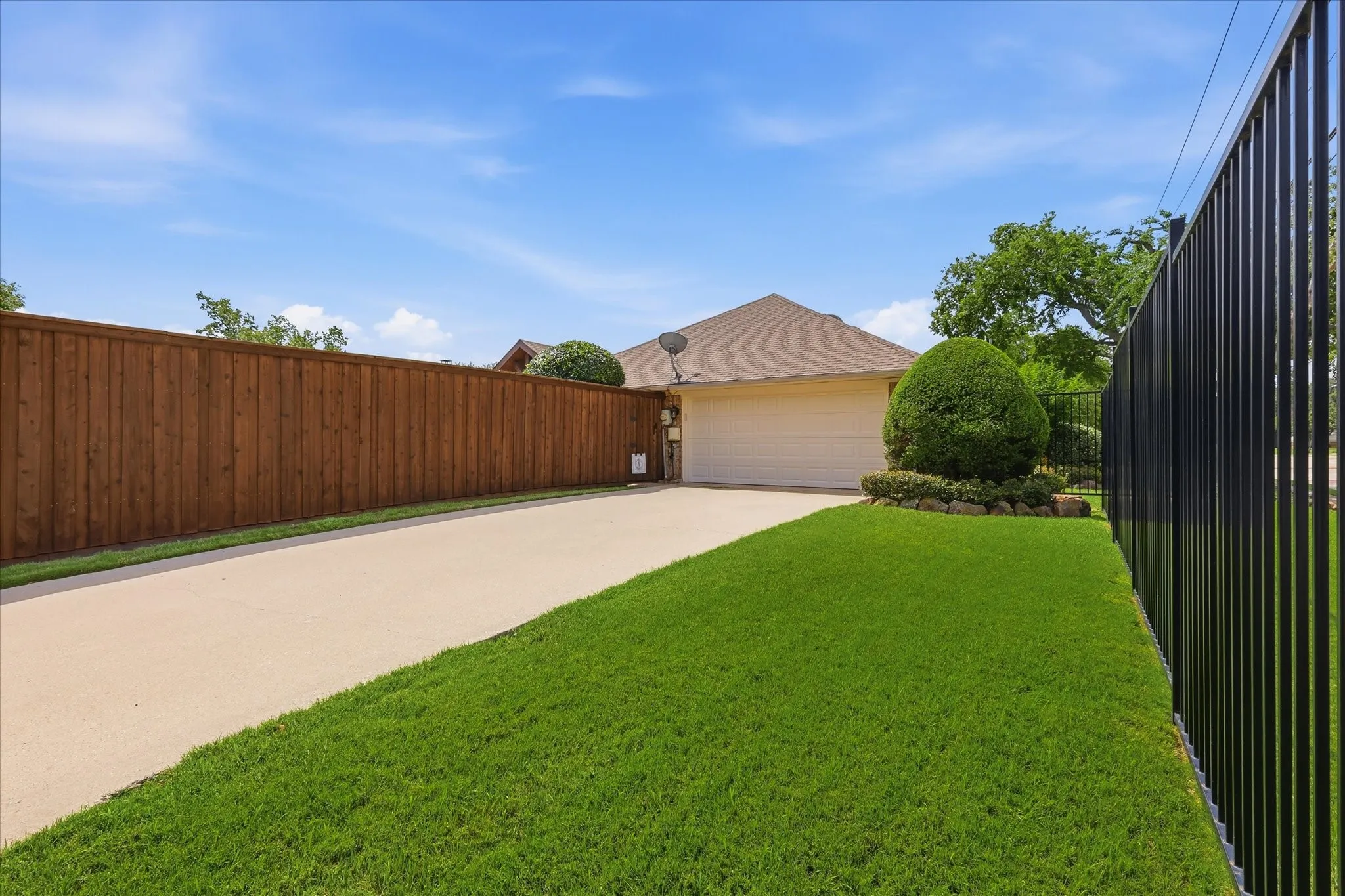 Spacious concrete driveway featuring a lush green lawn, full wood-plank fencing, and a black wrought-iron fence
