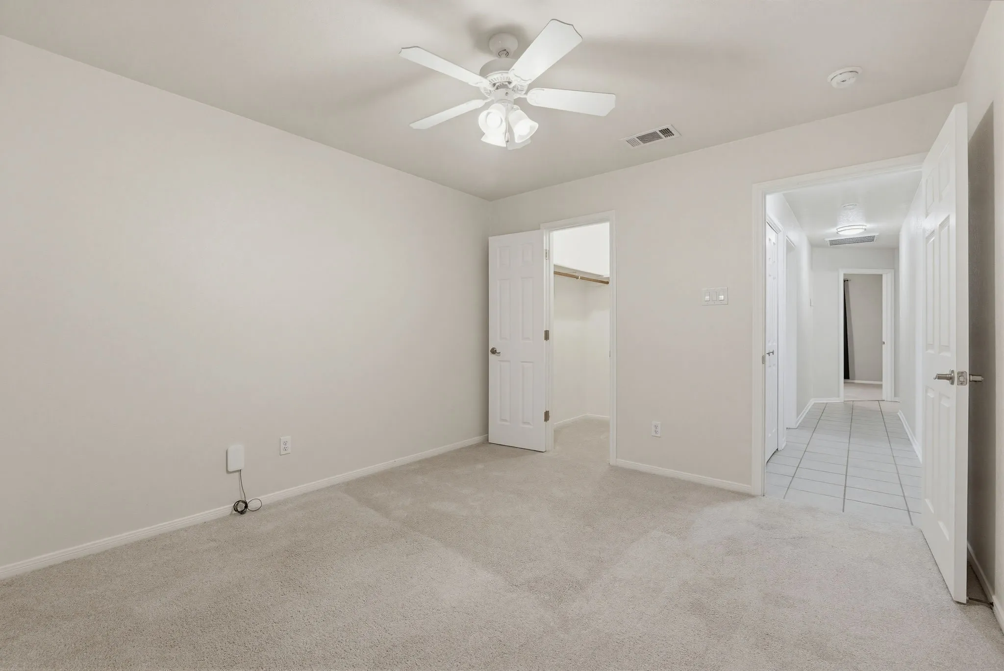 Carpeted room featuring a ceiling fan with light fixture, white walls, and a doorway opening to a hallway with tile flooring