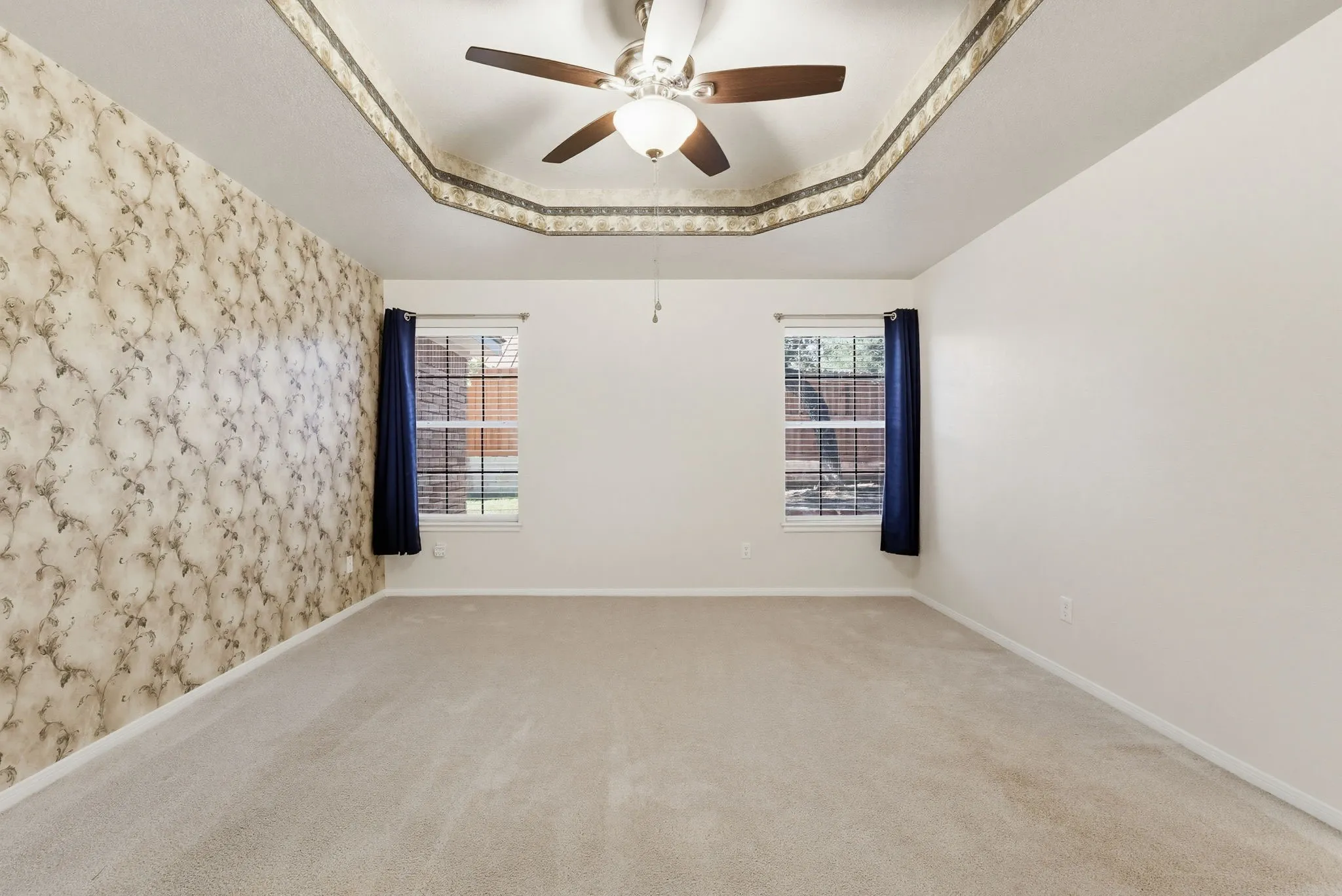 Spacious room featuring a tray ceiling with a ceiling fan, patterned accent wall, and two windows with blinds