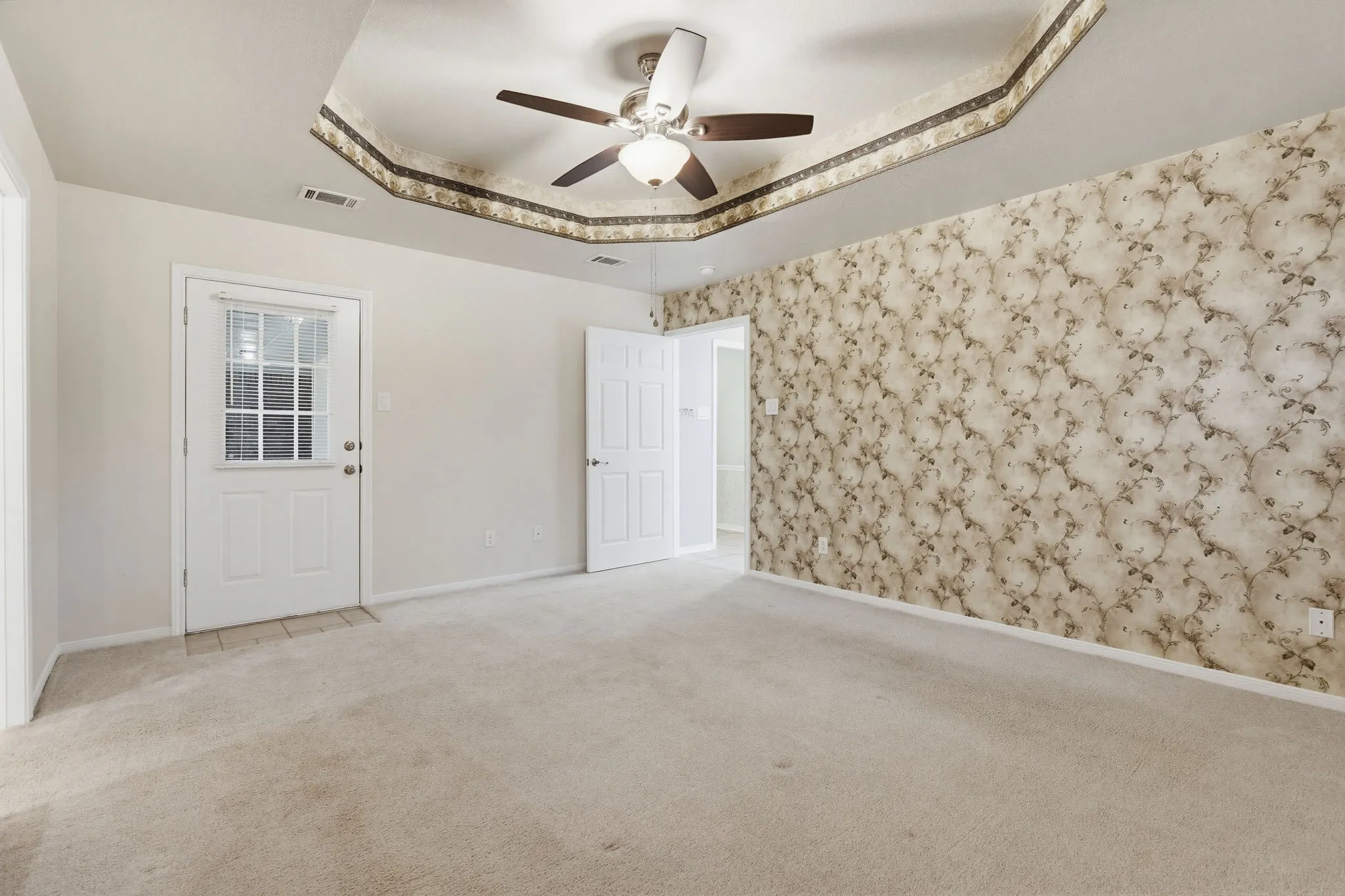 Spacious interior room featuring a tray ceiling with a ceiling fan, patterned accent wall, and light-colored carpeting