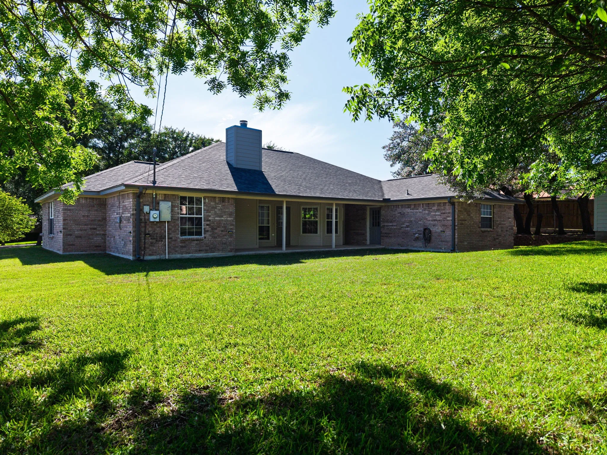 Expansive backyard featuring a lush lawn, mature trees, and a brick exterior home with a covered patio