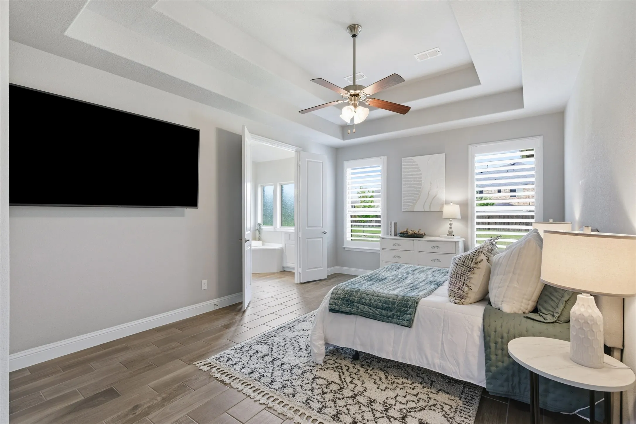 Spacious primary bedroom featuring wood-finish tile flooring, a tray ceiling with a ceiling fan, and white plantation shutters on multiple windows