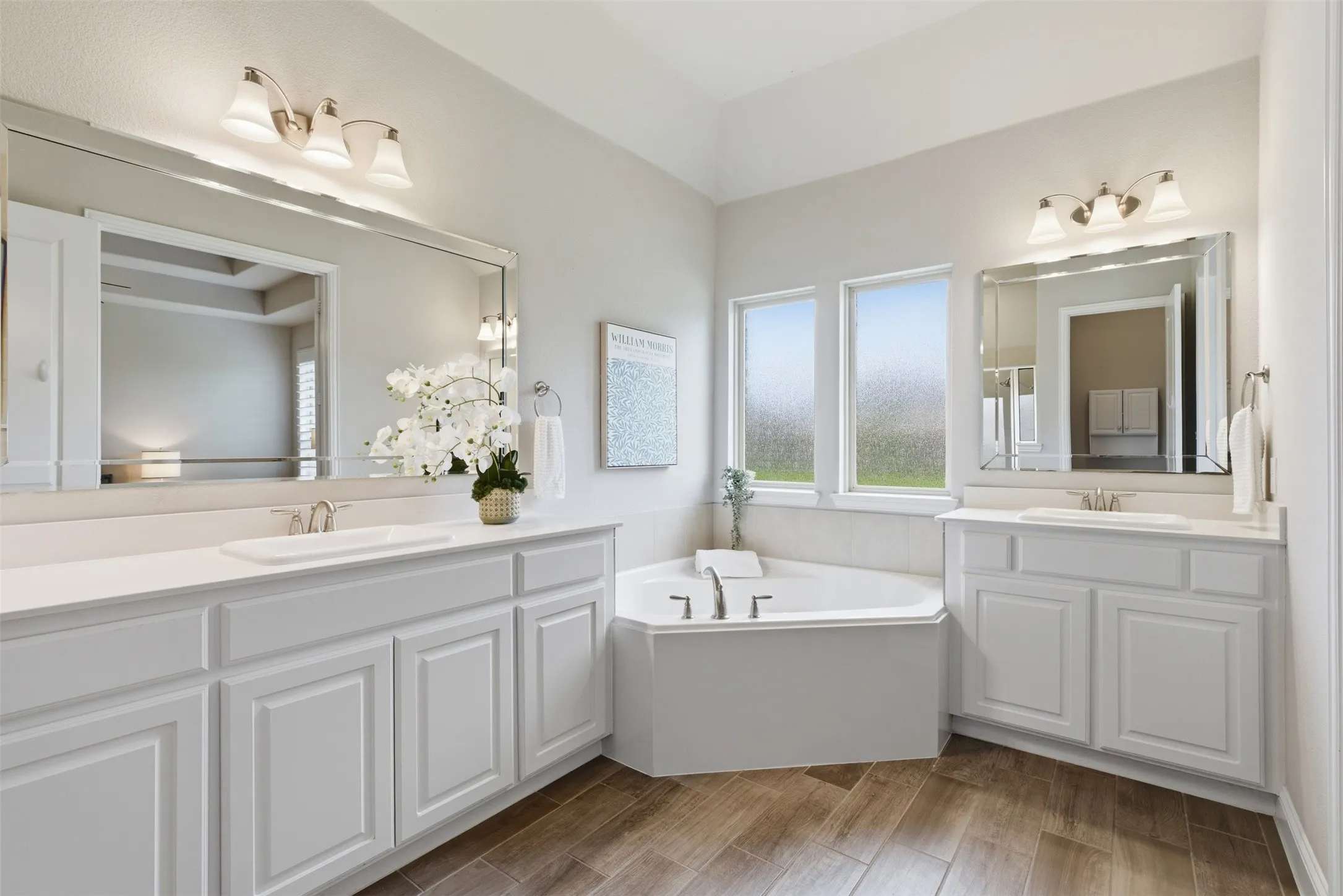 Bathroom featuring a corner soaking tub, dual white vanities with undermount sinks, and wood-finish flooring