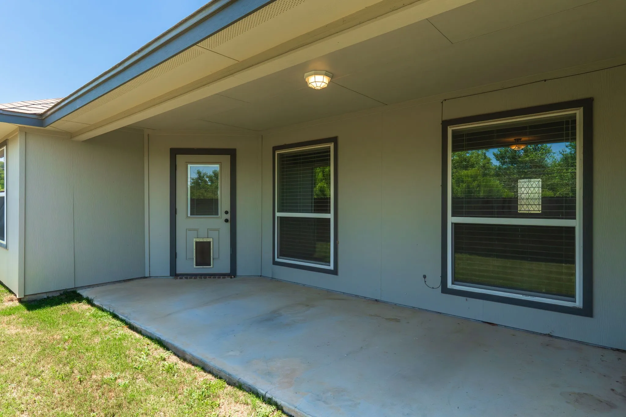 Covered concrete patio featuring an exterior door with a window insert, two large windows with blinds, a soffit, and an overhead light fixture