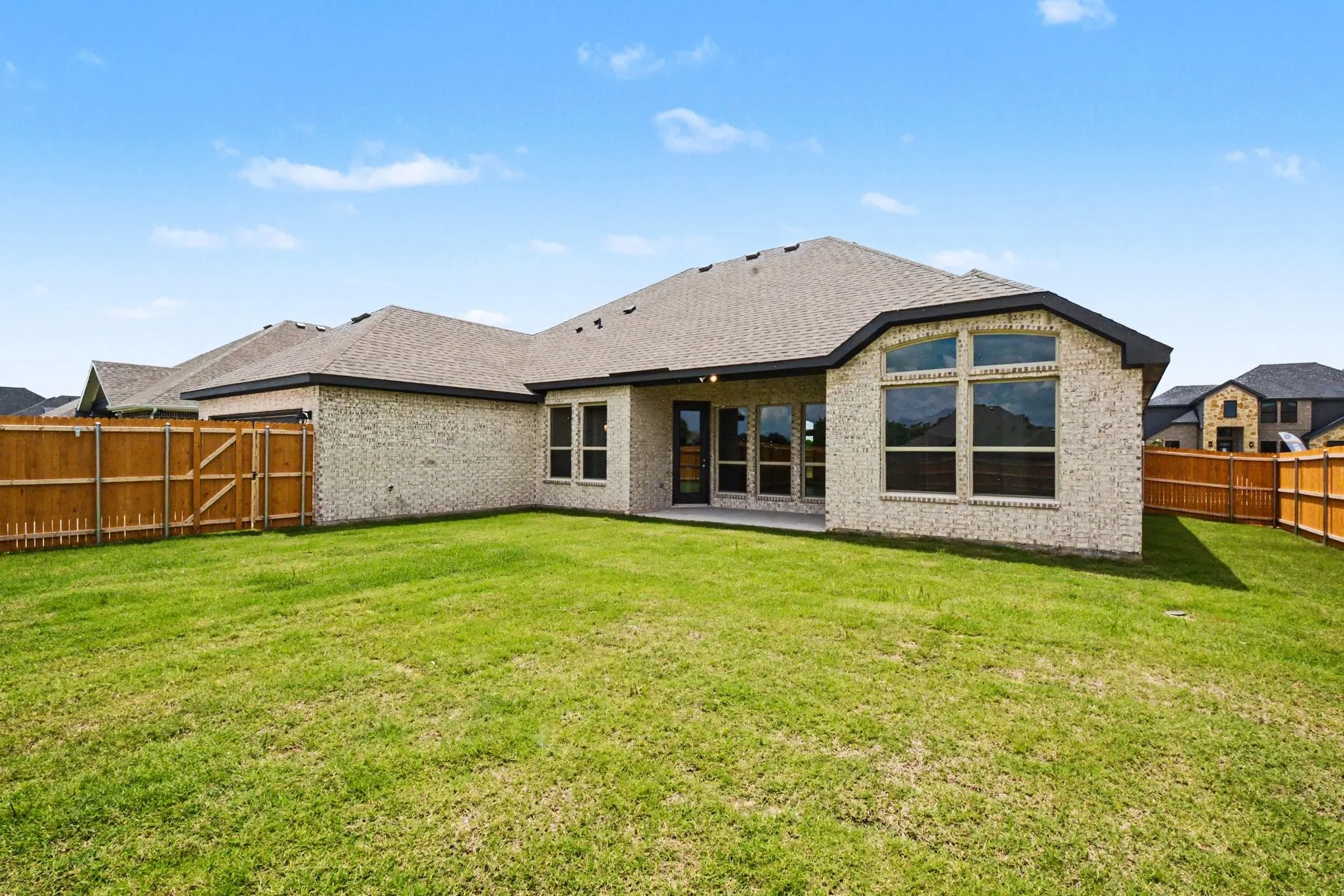 Expansive backyard featuring a manicured lawn, full wood fencing, a covered concrete patio, and a light-toned brick exterior with large windows